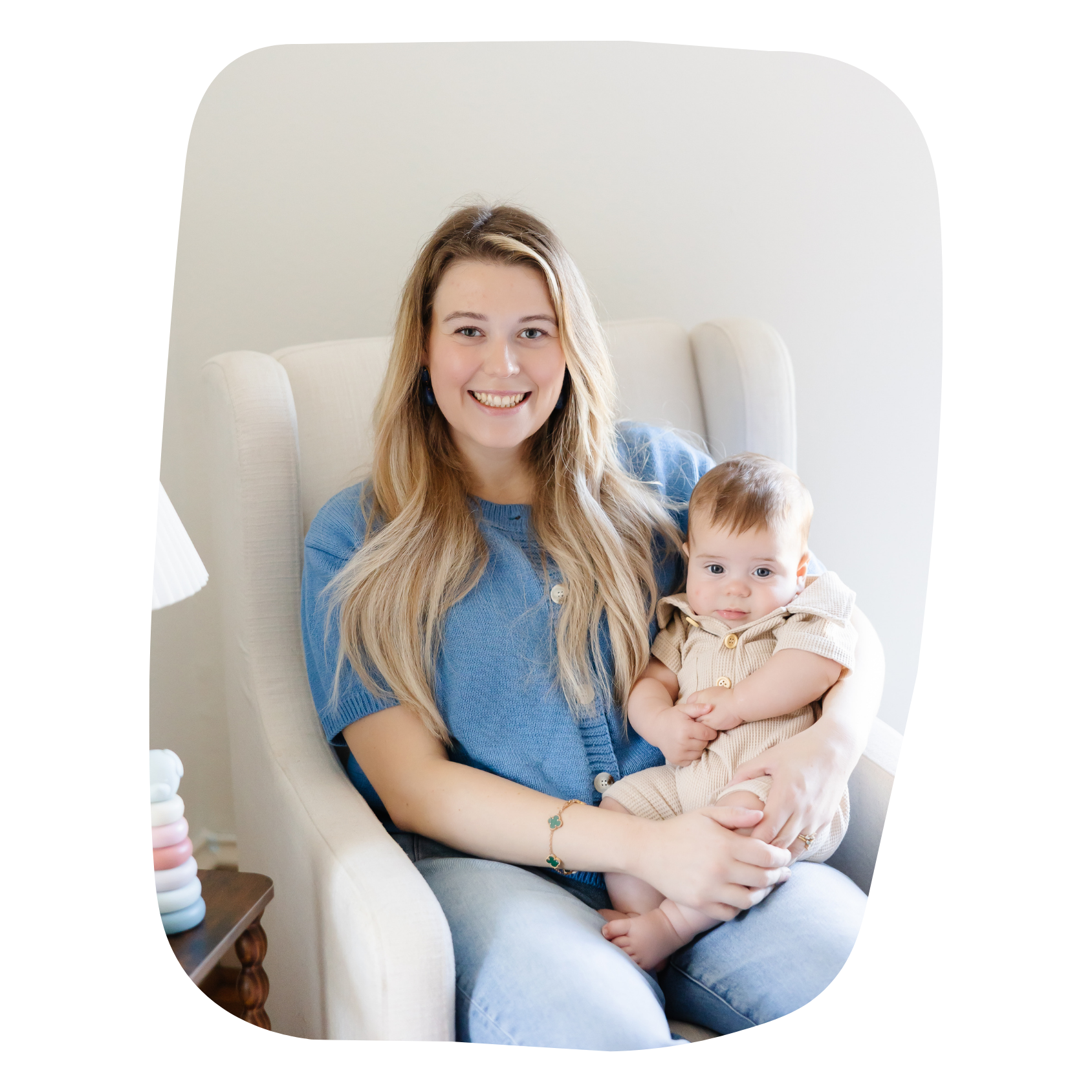 A smiling woman with long blonde hair sitting on a white armchair, holding a young child with light brown hair dressed in beige, in a cozy indoor setting.
