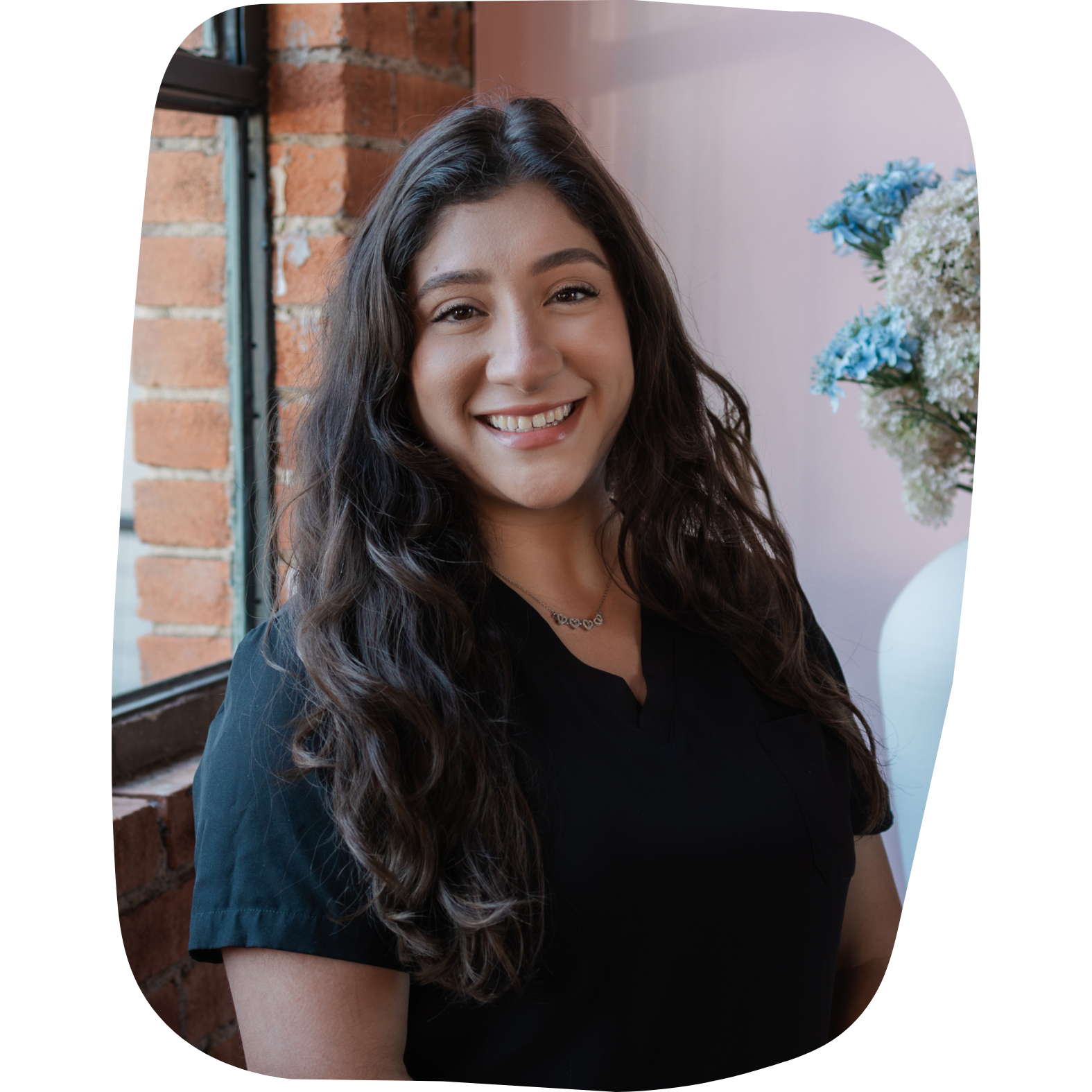A young woman with long, wavy dark hair smiling in front of a window with exposed brick and a pink wall, decorated with a vase of white and blue flowers.