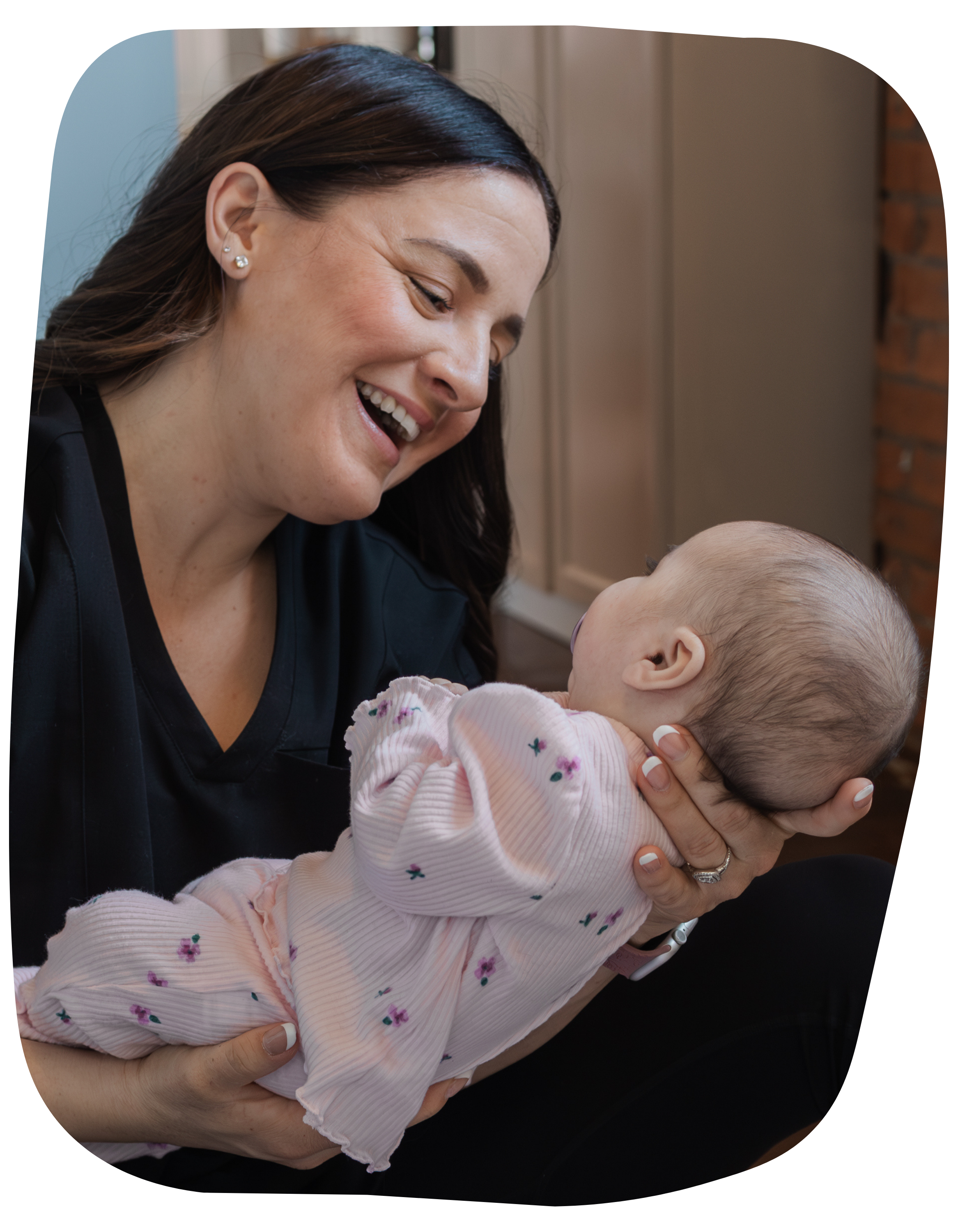 A woman smiling and holding a baby in her arms, looking at the baby with affection indoors.