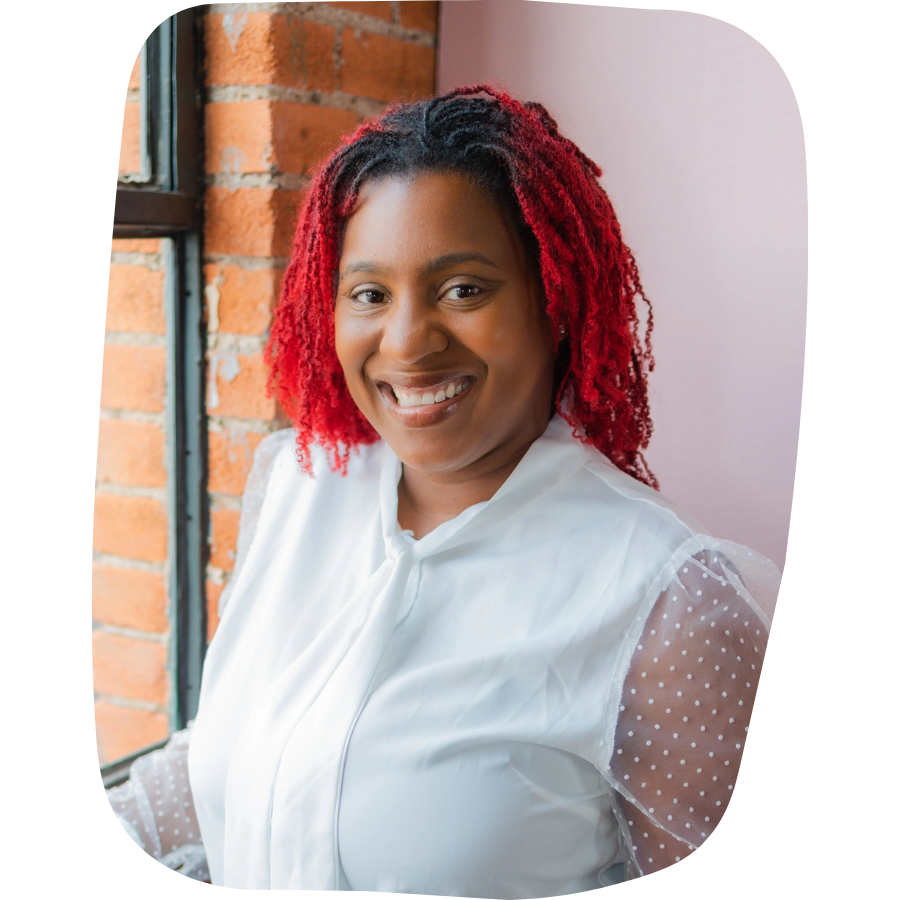 A smiling woman with red curly hair, wearing a white blouse with sheer polka-dot sleeves, standing near a window with brick wall in the background.