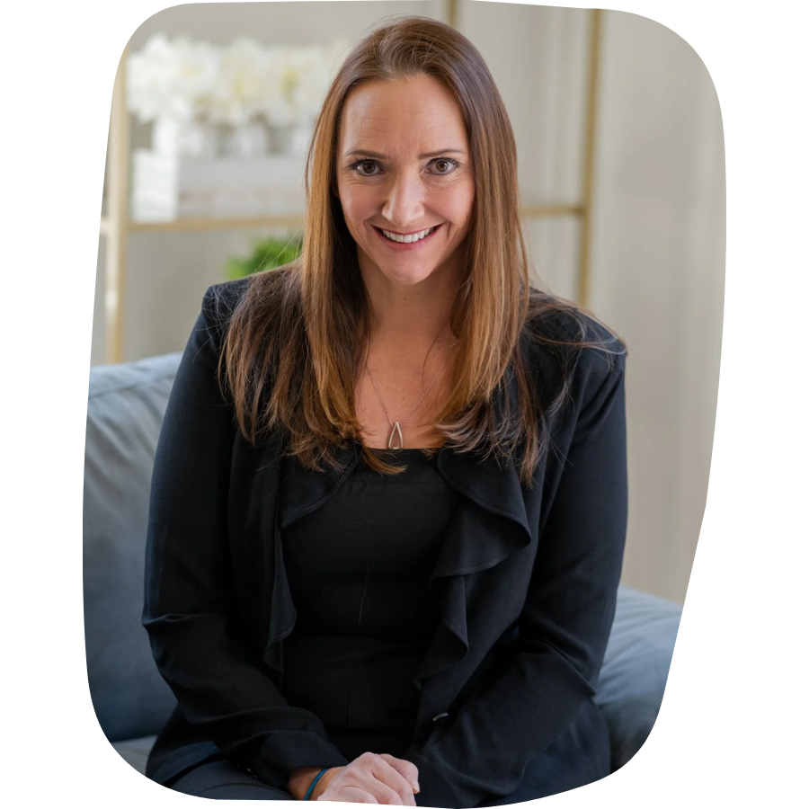 A woman with long brown hair smiling and sitting on a sofa in a well-lit room, wearing a black blazer and a necklace.