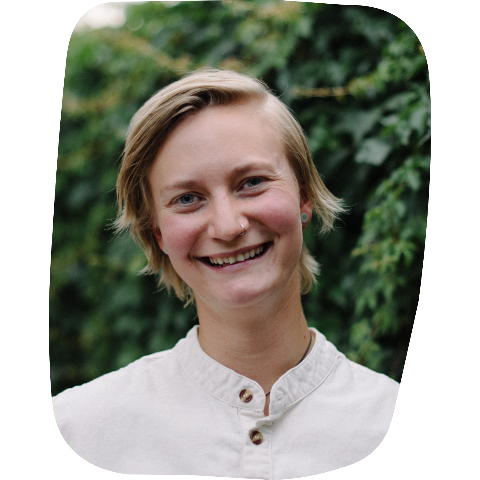 A young woman with short blonde hair, nose piercing, wearing a white button-up shirt, smiling outdoors with green foliage in the background.