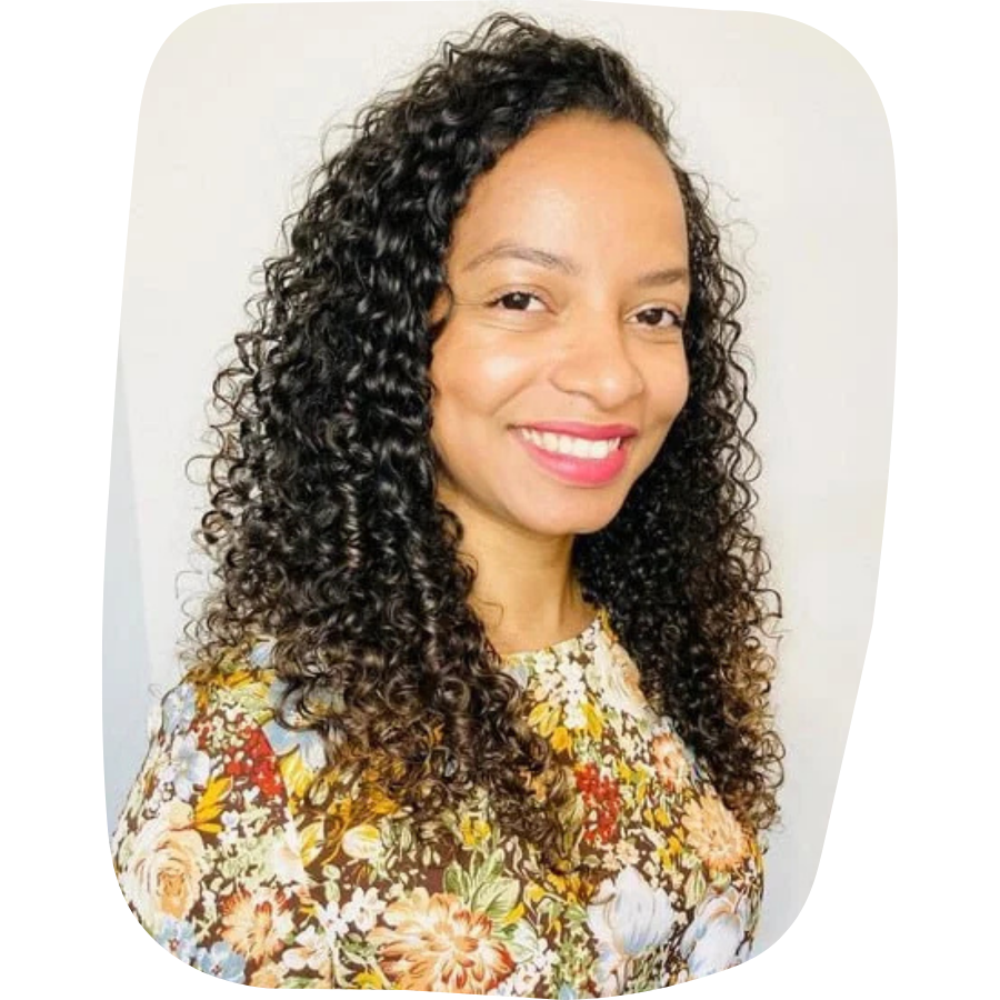 A woman with curly dark hair and a bright smile, wearing a floral blouse, standing against a plain light background.