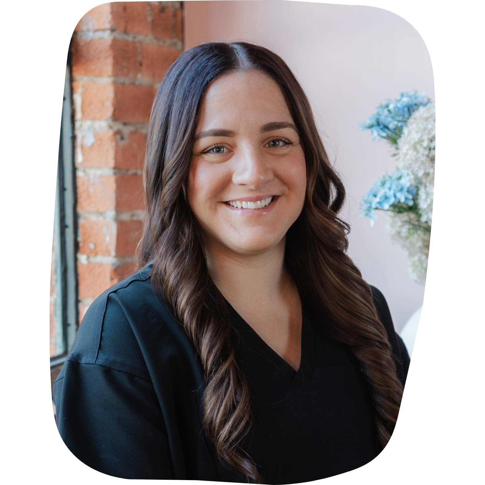 A smiling woman with long brown hair wearing a black top, sitting indoors near a brick wall and a window with flowers in the background.