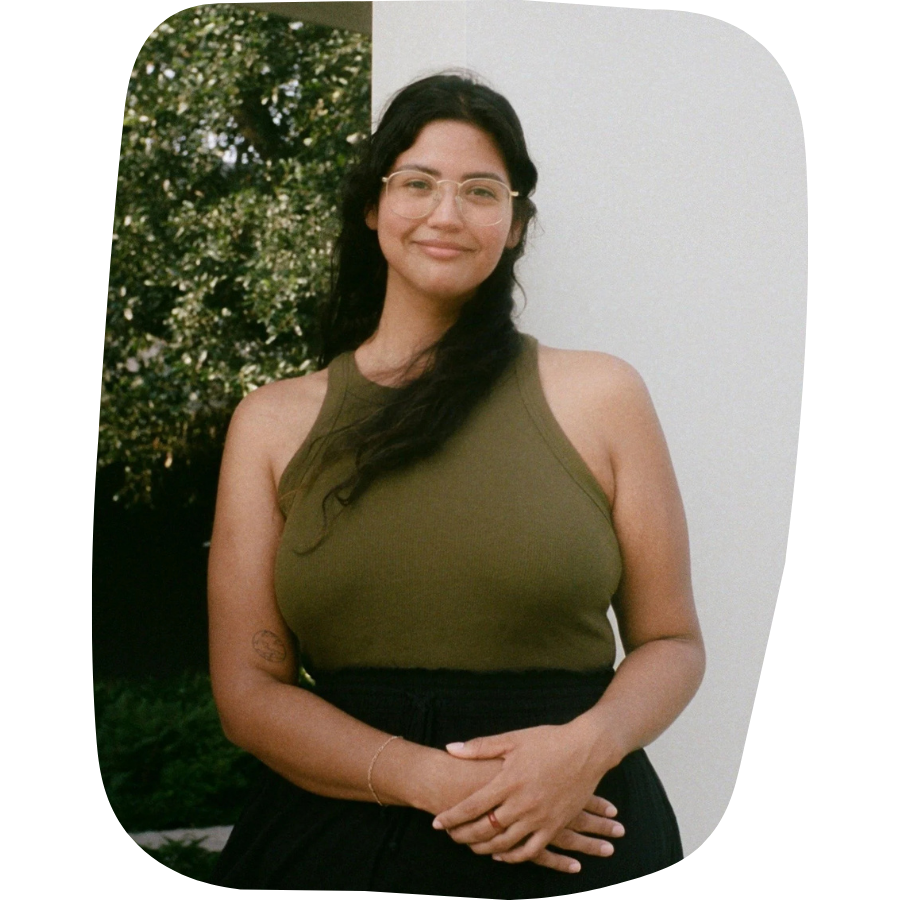 A woman with long dark hair, glasses, and a slight smile, standing outdoors in front of greenery and a white wall, wearing an olive green sleeveless top and black bottoms.