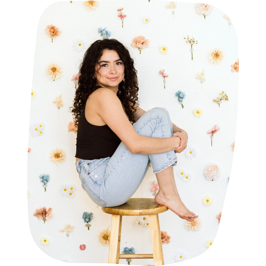 A young woman with curly dark hair sitting on a wooden stool, her knees drawn up to her chest, against a background decorated with colorful flowers.