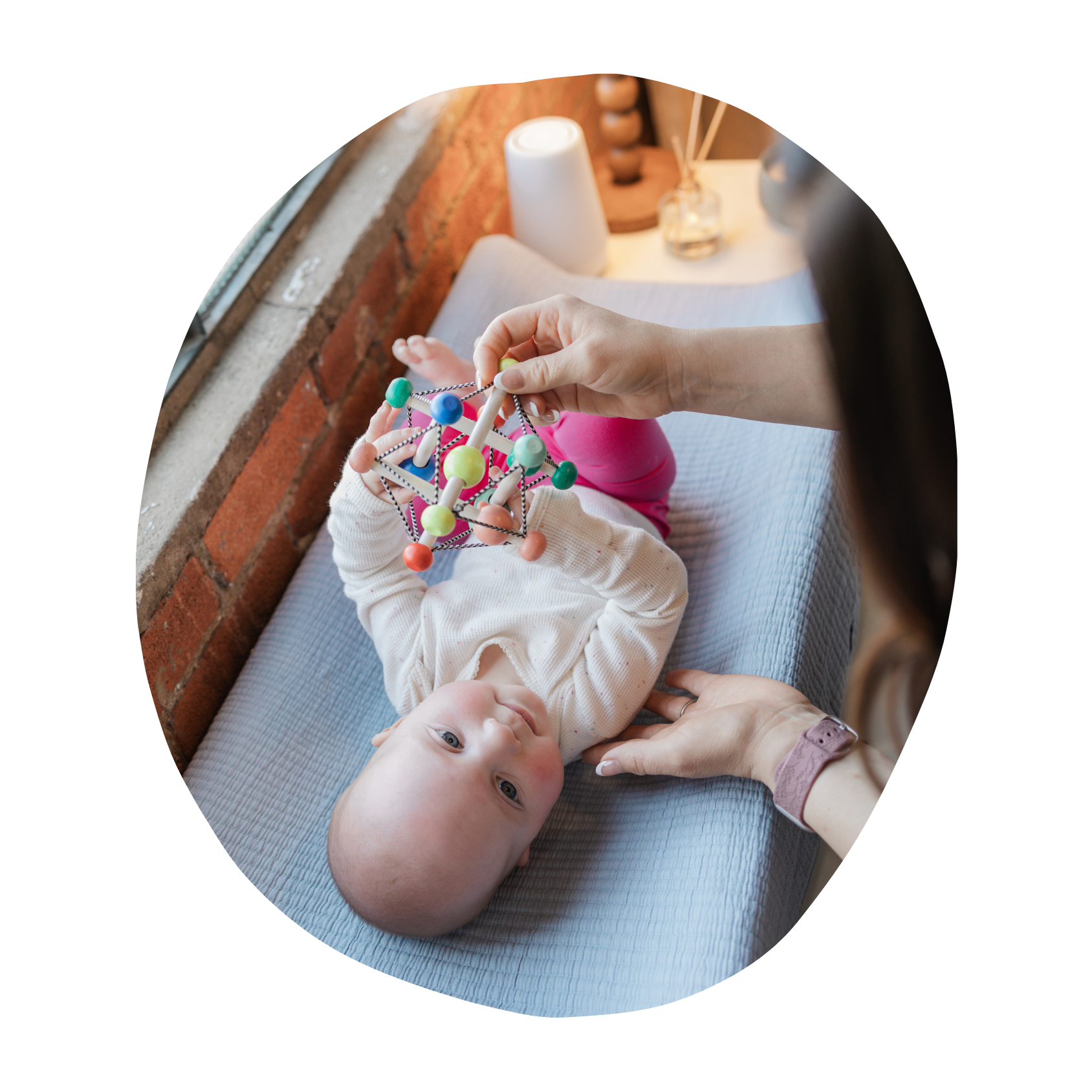 Child lying on bed sharing a colorful toy with an adult, with brick wall, window, and personal items on side table in background.