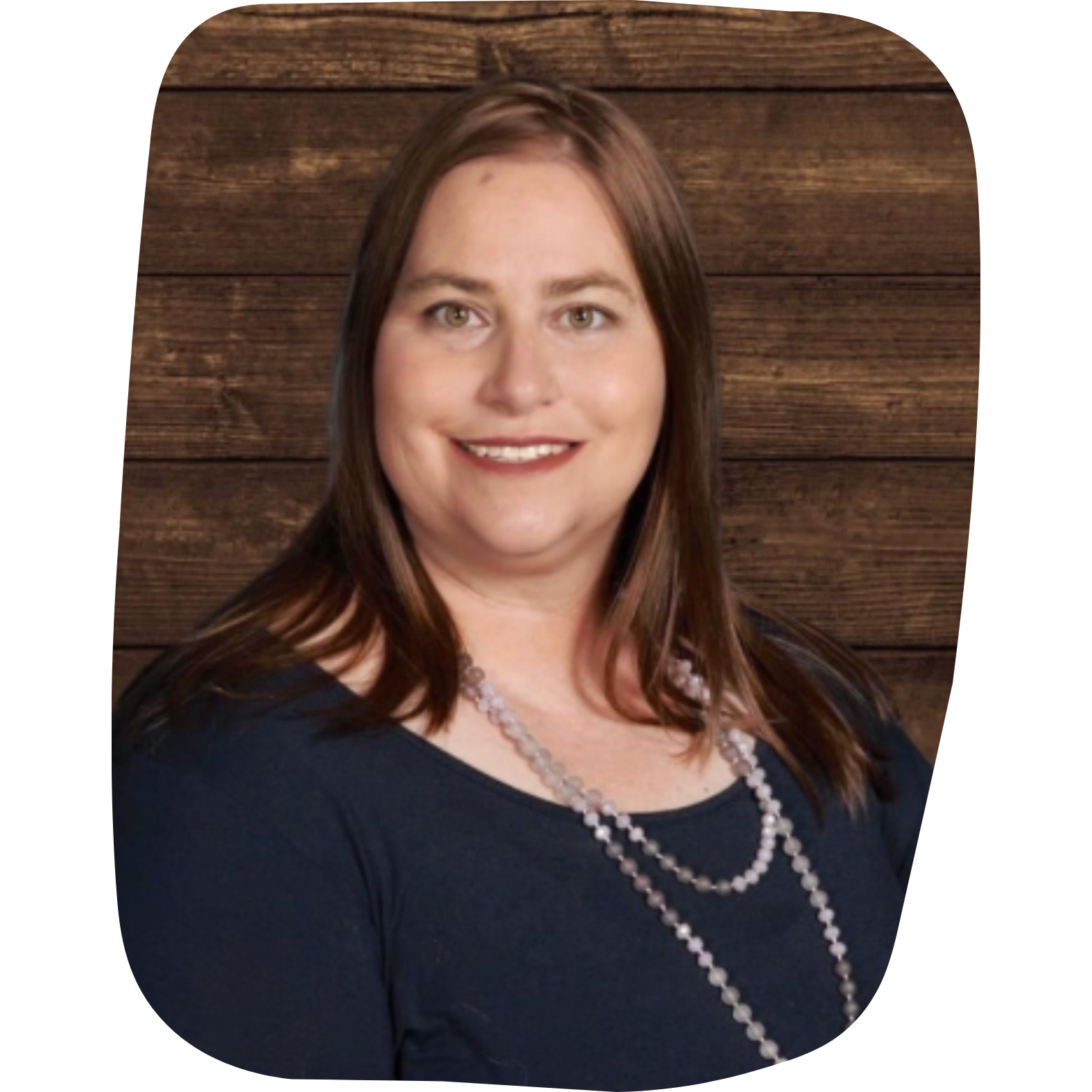 Portrait of a woman with brown hair wearing a navy blue top and a pearl necklace, standing in front of a wooden background.
