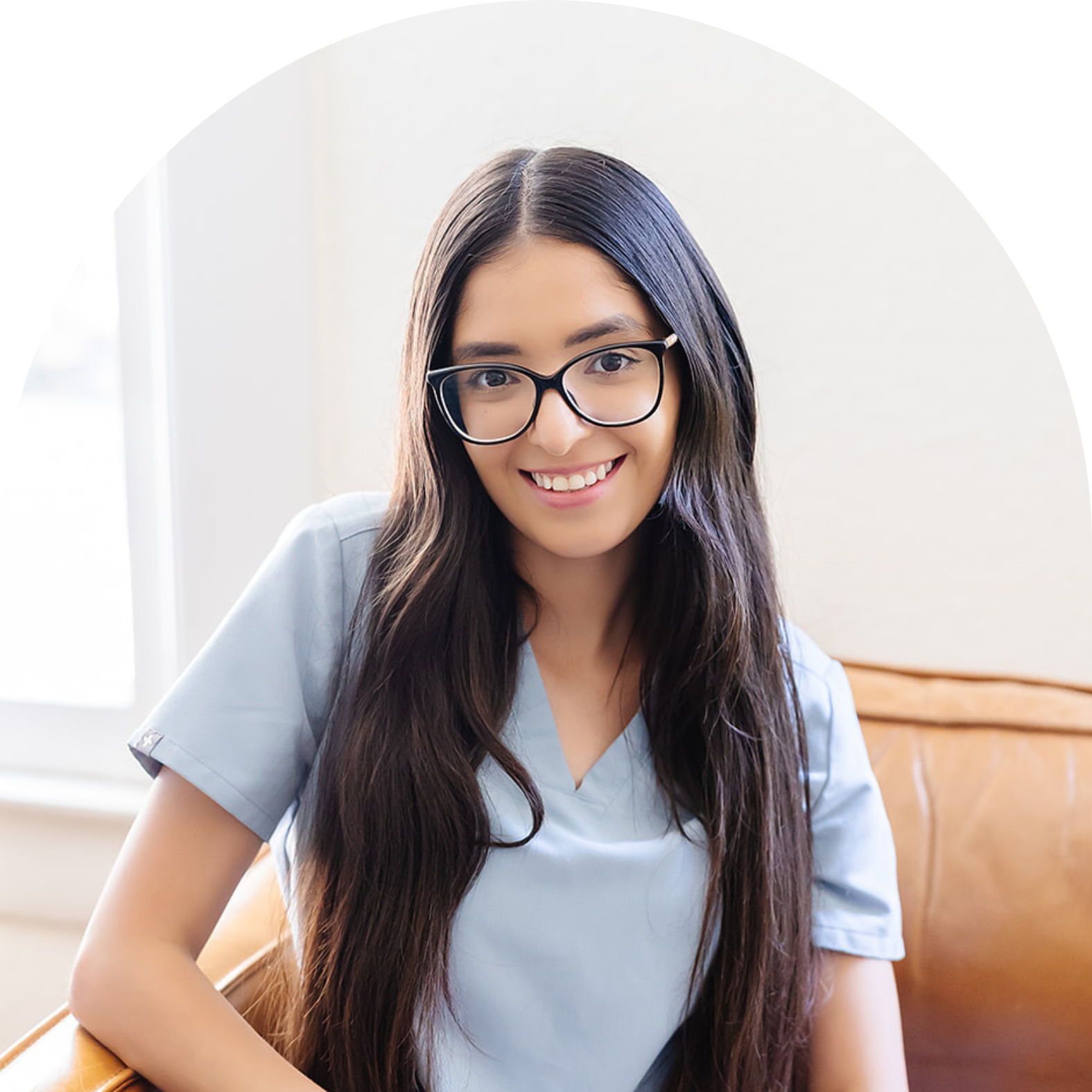 A young woman with long dark hair, glasses, and a light blue medical scrub top, smiling while sitting on a brown leather couch.