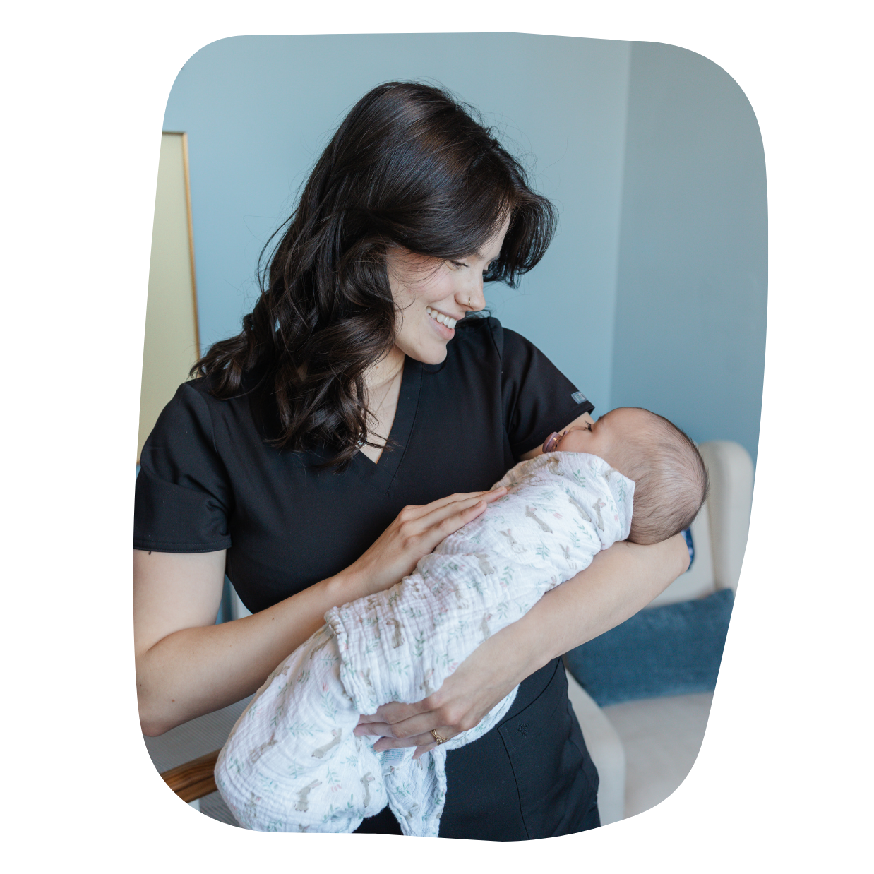 A young woman holding a smiling baby in her arms, both indoors. The woman has dark, wavy hair and is wearing a black top. The baby is wrapped in a light-colored patterned blanket.