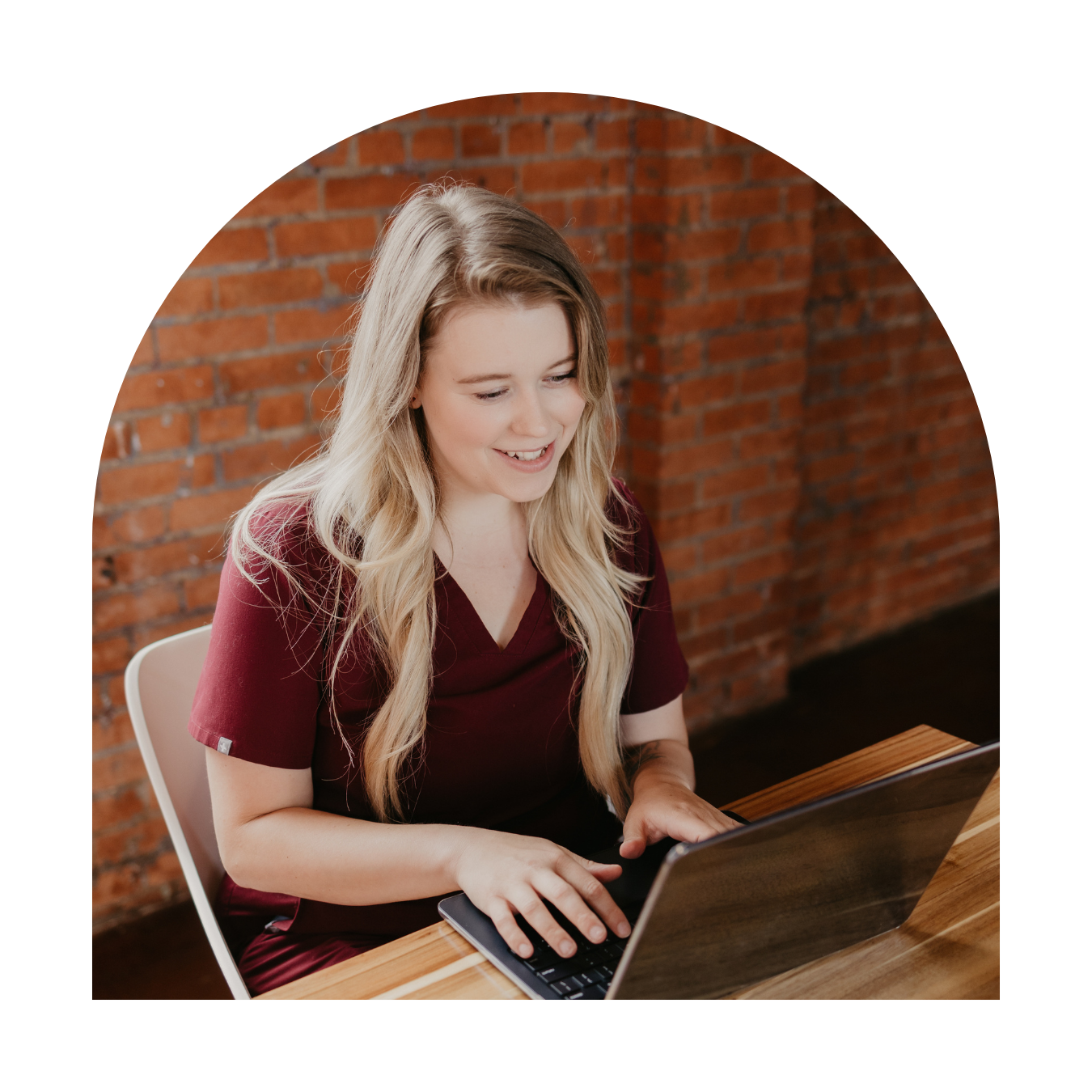 Smiling young woman with blonde hair working on a laptop at a wooden table in front of a brick wall.