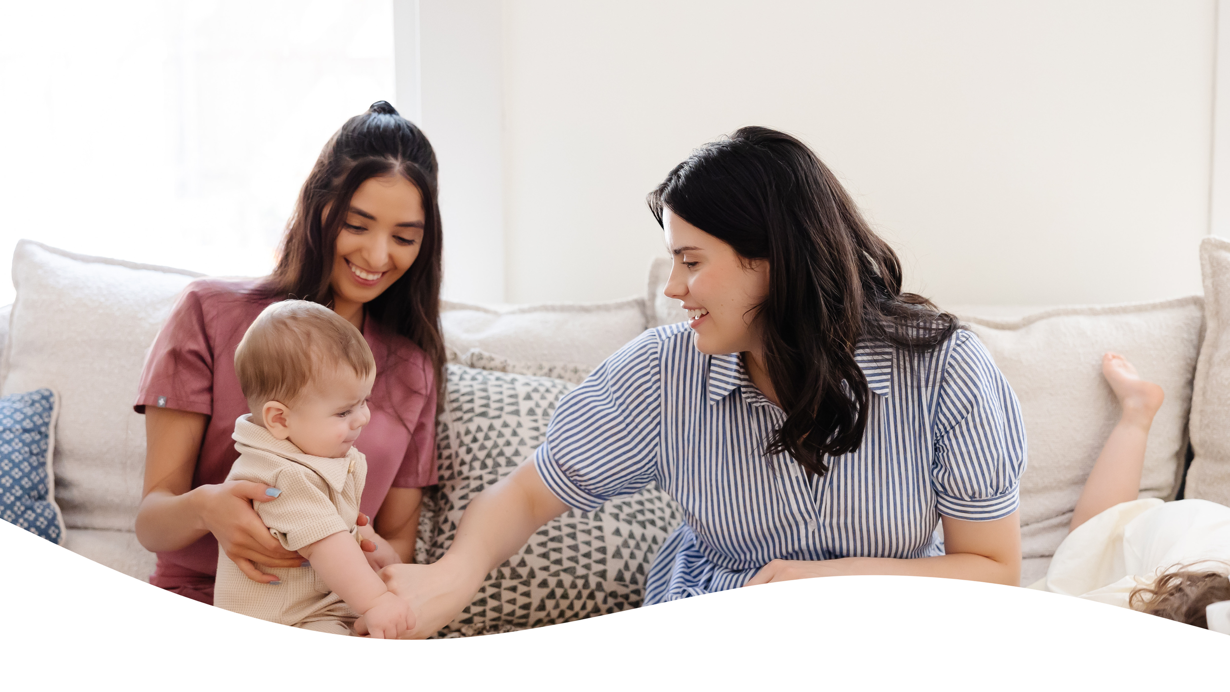 Two women and a young child sitting on a sofa, smiling and playing together in a bright living room.