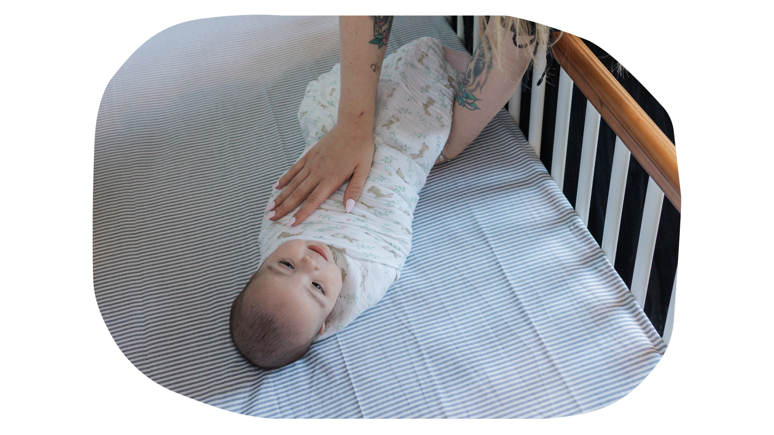 A baby lying on a bed with a striped bedsheet, receiving gentle abdominal massage from an adult with tattooed arms.