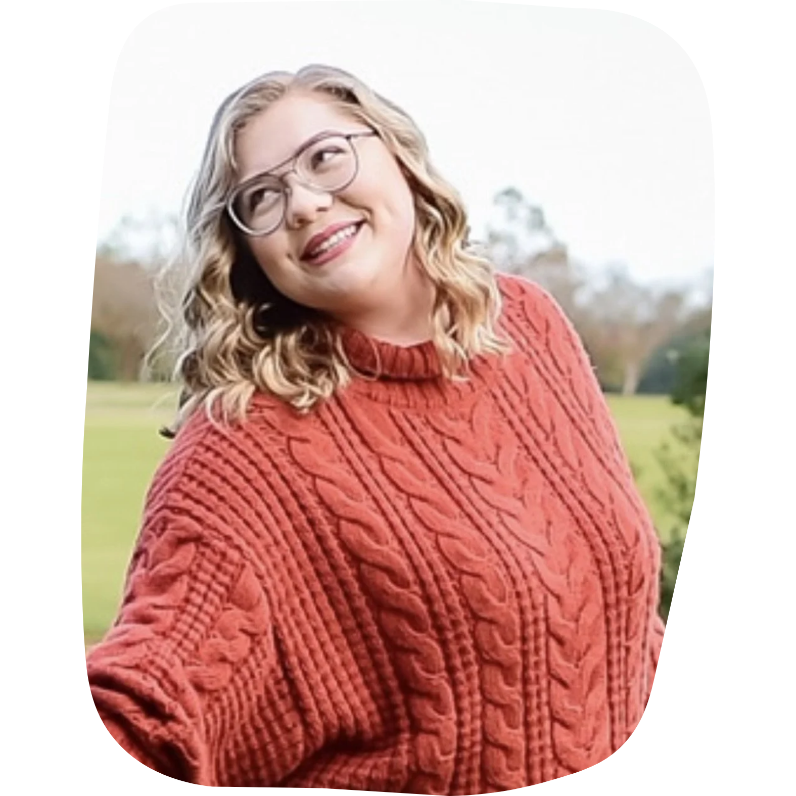 A young woman with glasses and curly blonde hair wearing a rust-colored cable knit sweater, outdoors in a park during autumn.