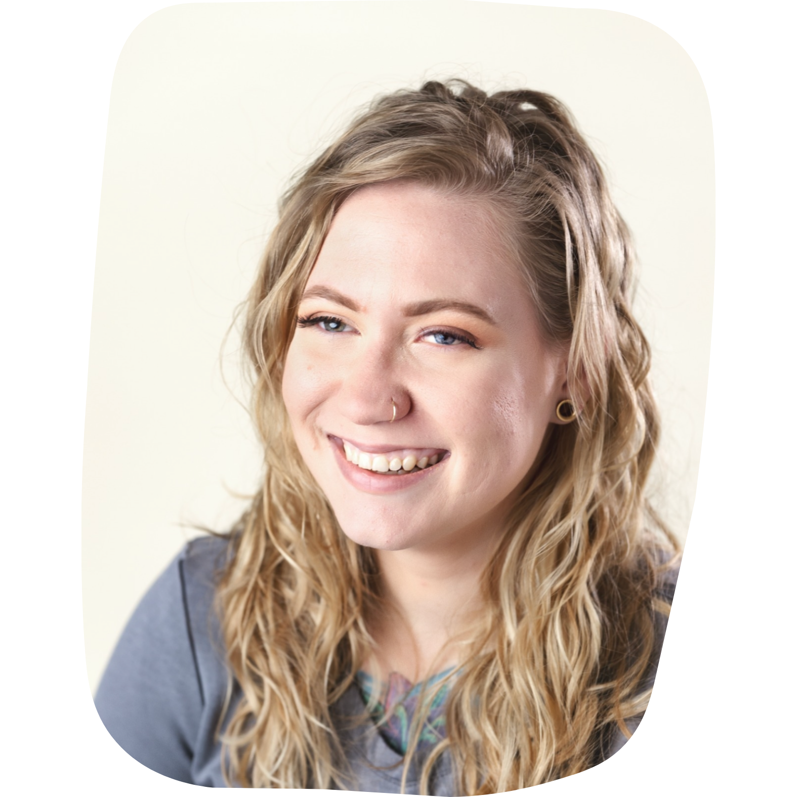 Close-up of a young woman with wavy blonde hair smiling, wearing a nose ring and earrings, against a neutral background.