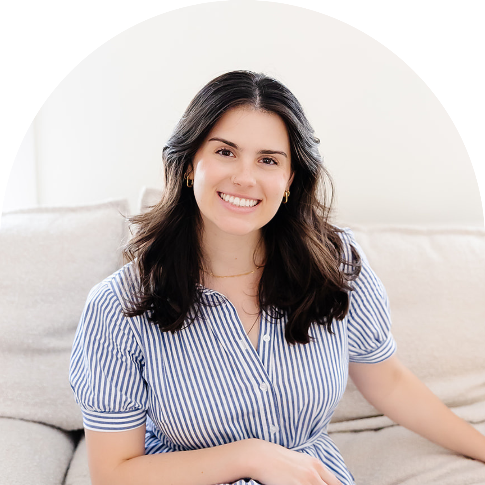 A woman with long dark hair smiling and sitting on a beige sofa in a bright room, wearing a blue and white striped shirt and gold jewelry.