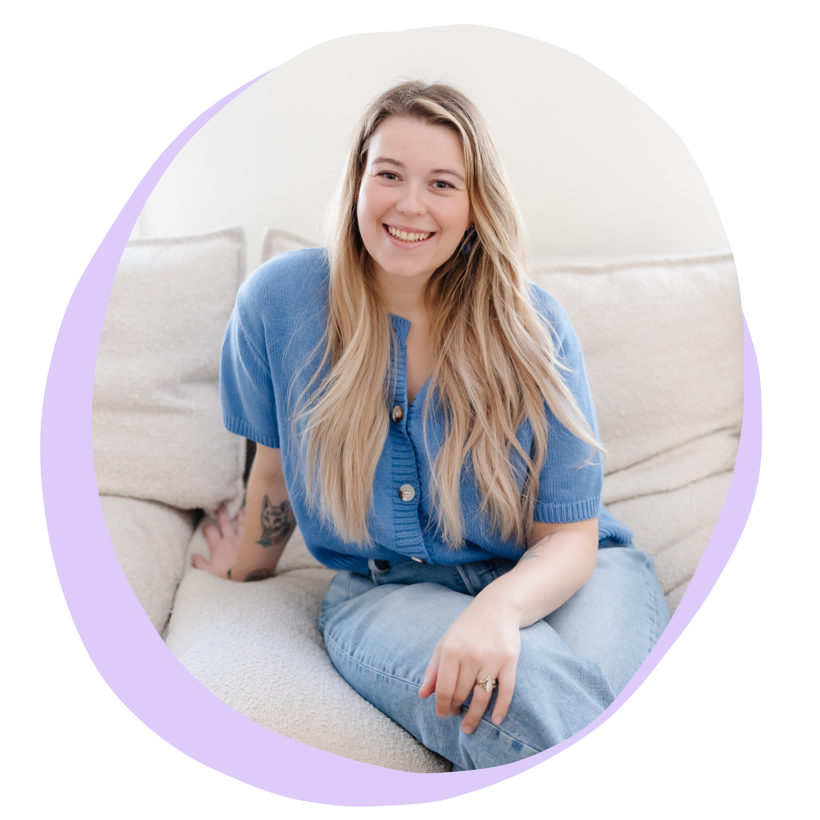 A young woman with long blonde hair, wearing a blue cardigan and jeans, sitting on a beige couch and smiling at the camera.