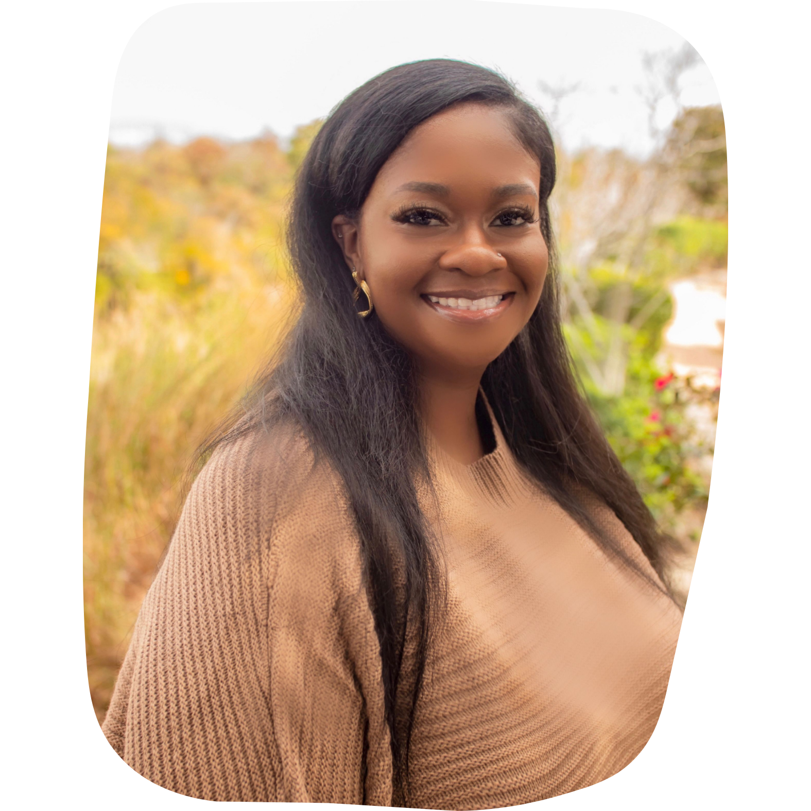A woman smiling outdoors with a blurred natural background, wearing a beige sweater and gold earrings.