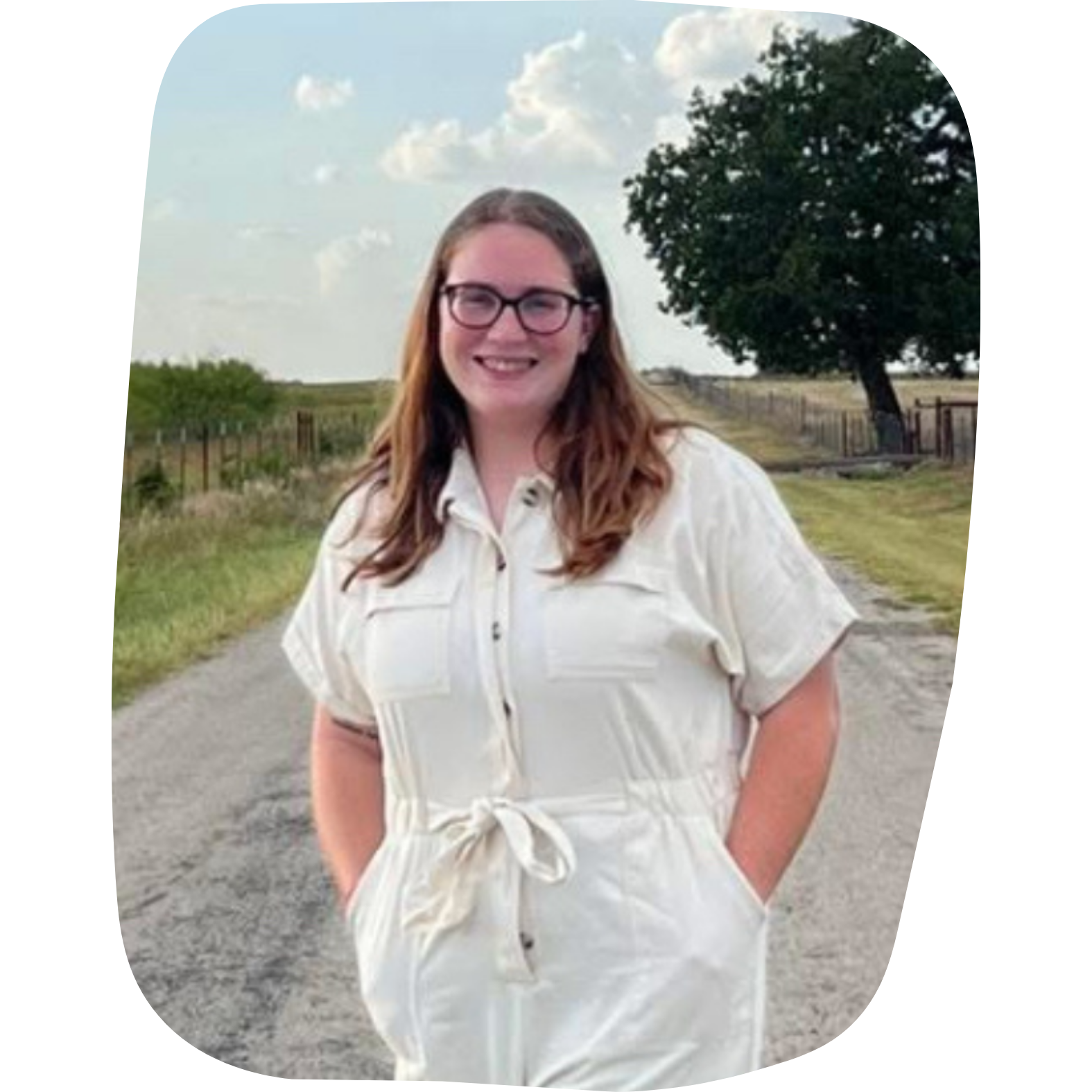 A smiling woman with glasses and long reddish-brown hair standing outdoors on a rural path with fields, trees, a fence, and a partly cloudy sky in the background.