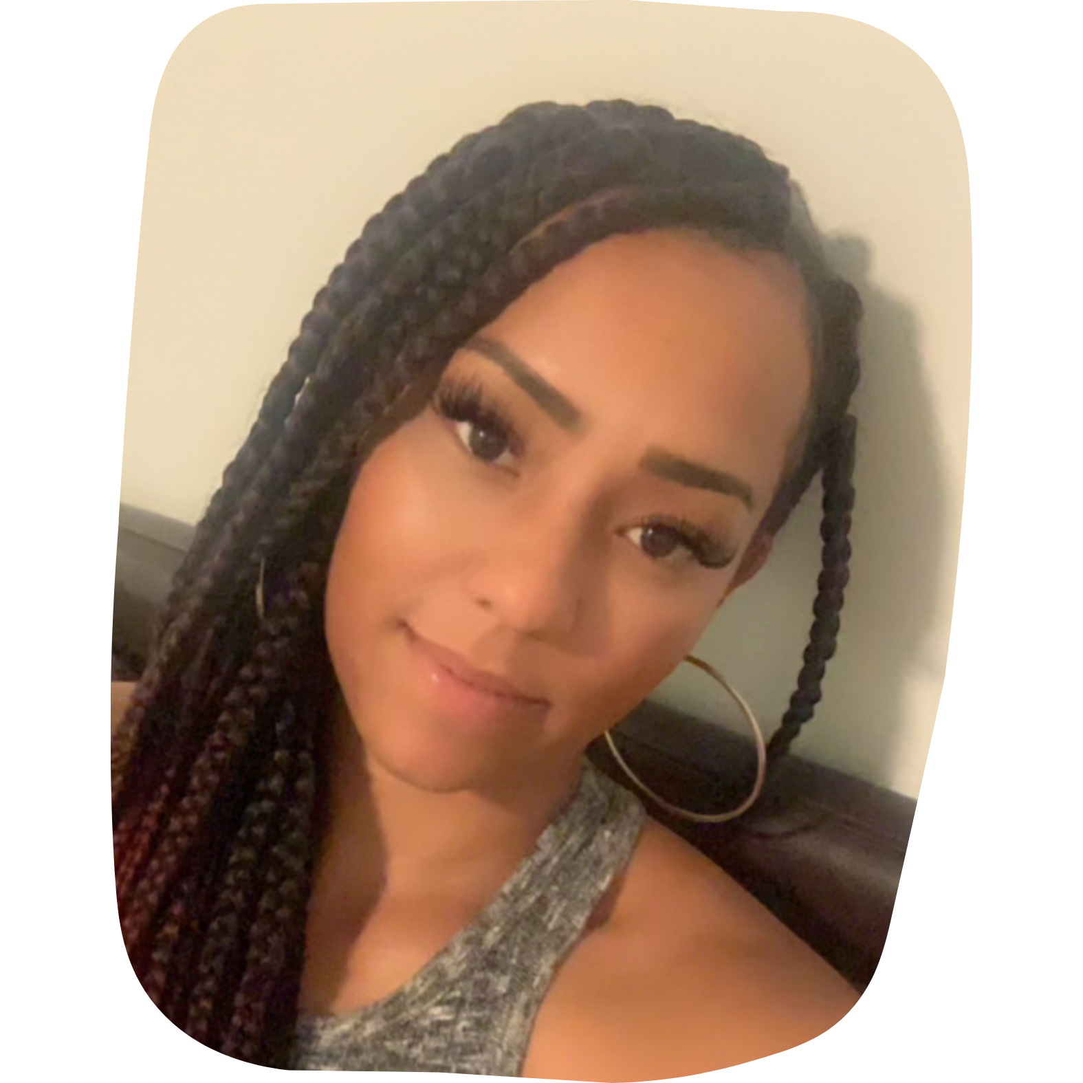A young woman with long braided hair, hoop earrings, and makeup, smiling and looking at the camera indoors.