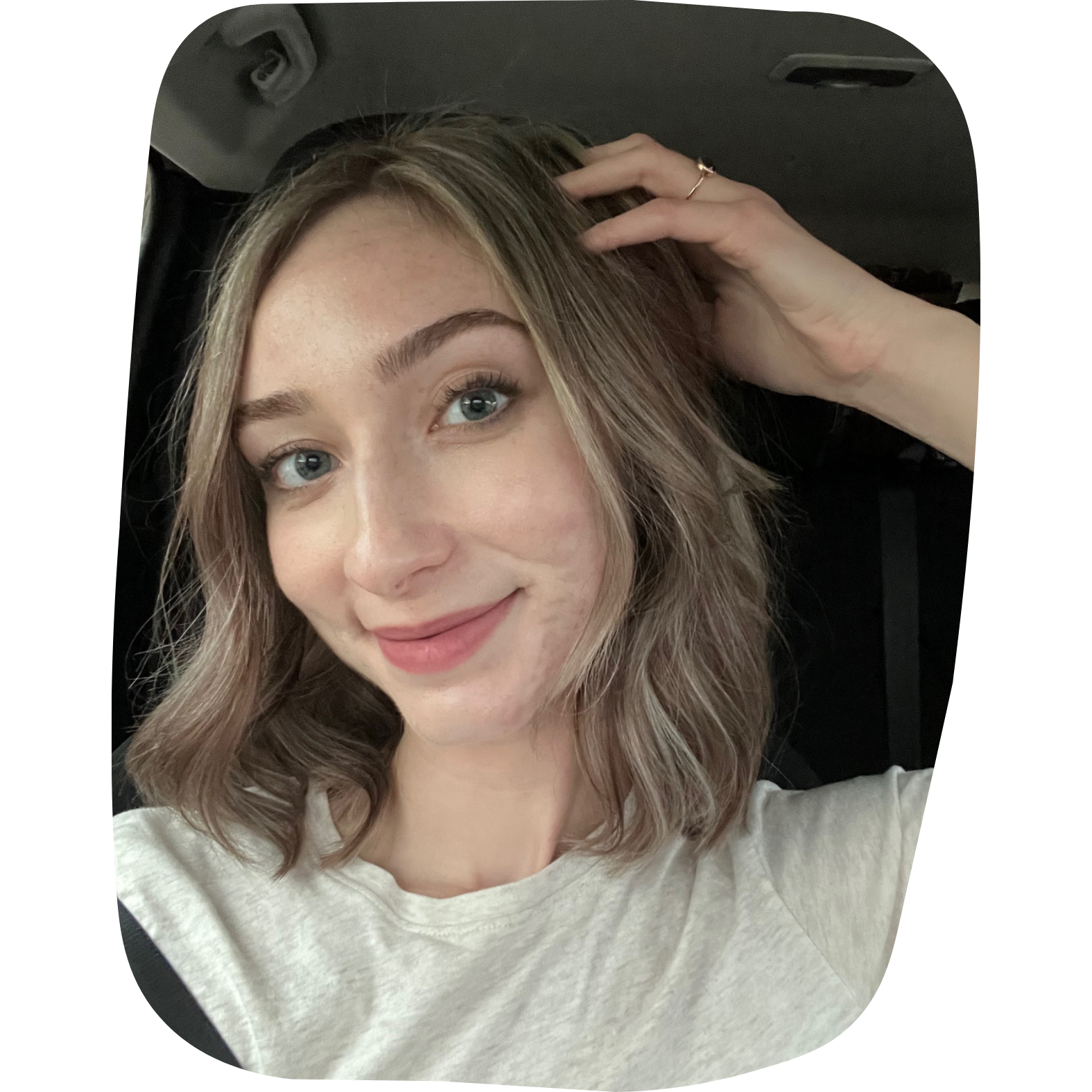 A young woman with light brown, wavy hair and blue eyes smiling at the camera inside a vehicle, wearing a light-colored t-shirt.