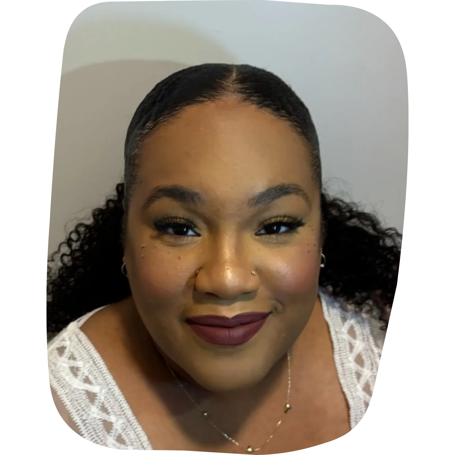 Close-up of a woman smiling with dark lipstick, makeup, and wearing a white lace top. She has dark curly hair and wears jewelry including earrings, a nose ring, and a necklace.
