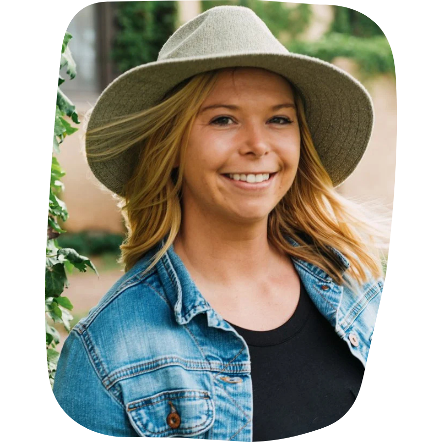 A smiling young woman with blonde hair wearing a wide-brimmed gray hat, a black shirt, and a denim jacket outdoors.