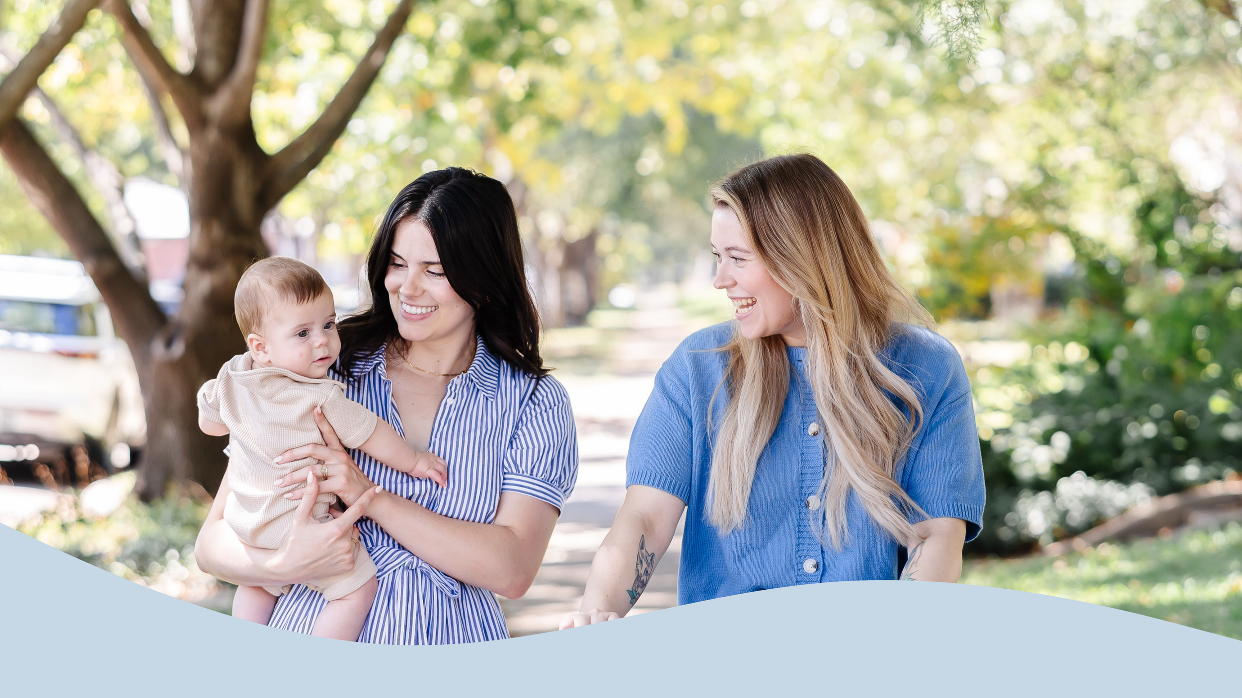 Two women, one with dark hair and one with blonde hair, smiling and talking while walking outdoors with a baby girl who has light brown hair, in a park setting with trees and greenery.
