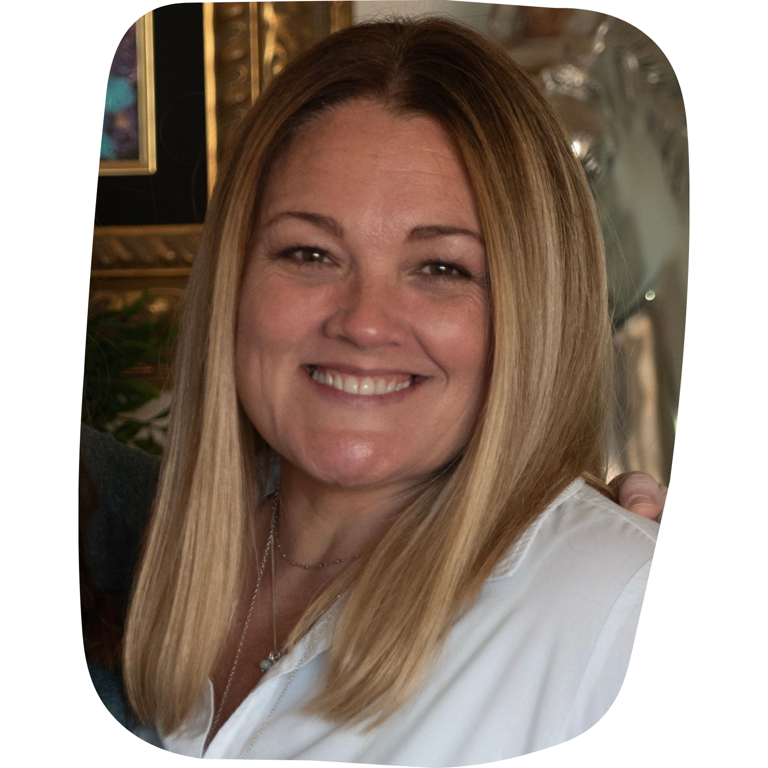 Close-up of a smiling woman with long blonde hair, wearing a white shirt and jewelry, in a warmly decorated interior setting.
