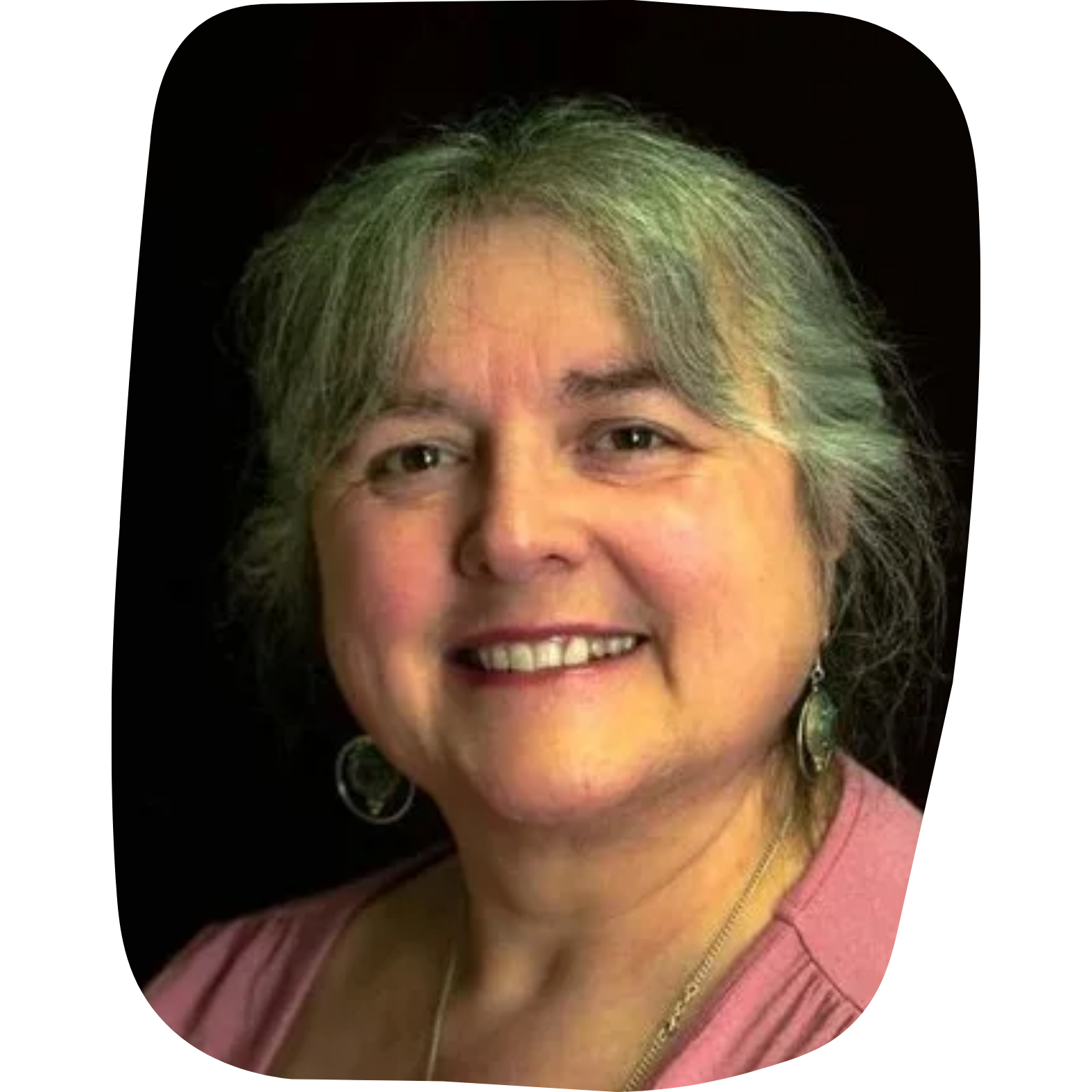 A smiling woman with short, grayish hair, earrings, wearing a pink top, against a black background.