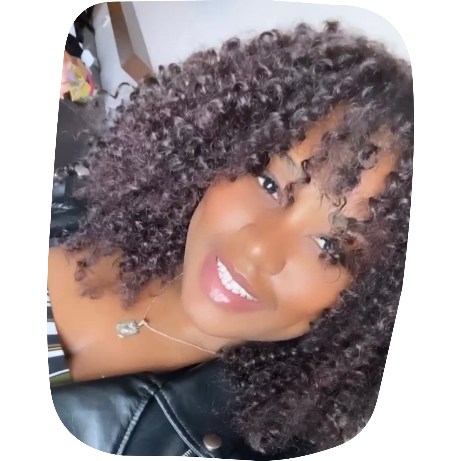 A woman with dark curly hair, smiling, wearing a necklace and black leather jacket.