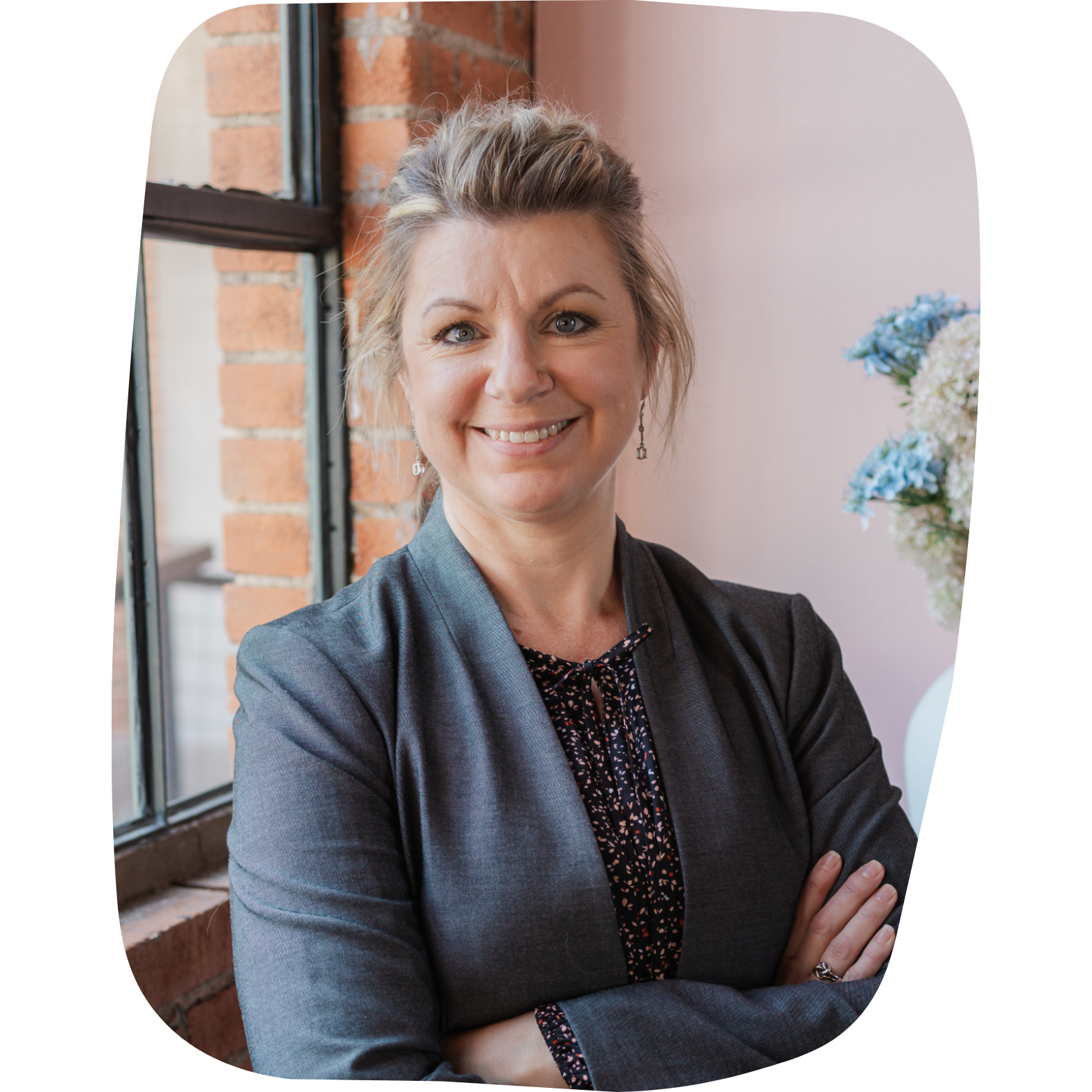A woman with blonde hair smiling, wearing a dark gray blazer and earrings, standing in front of a brick wall and a window with flowers in the background.