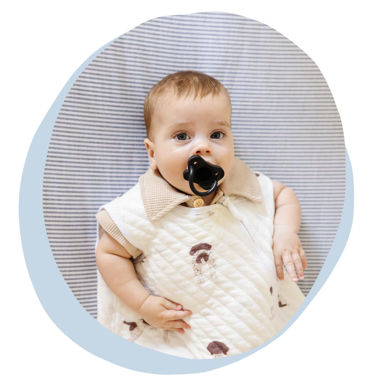 A young child with a pacifier in his mouth, lying on a pillow with a striped cover, wearing a beige vest with mushroom patterns.