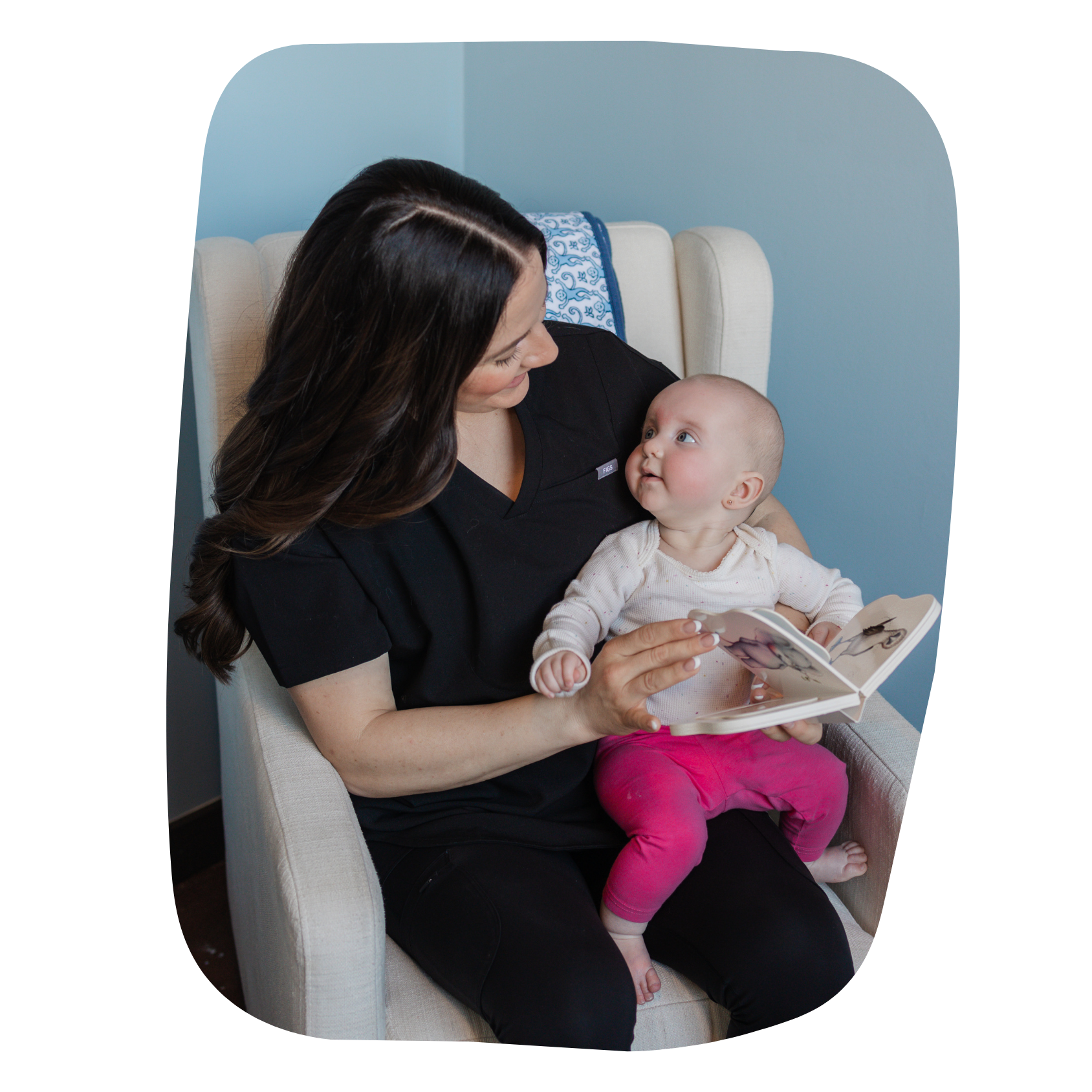A woman holding a baby girl on her lap while reading a picture book together on a beige armchair in a room with light blue walls.