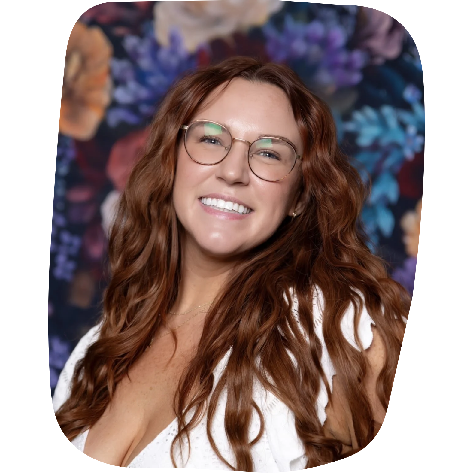 A woman with long red wavy hair and glasses smiling, wearing a white top, with a colorful floral background.