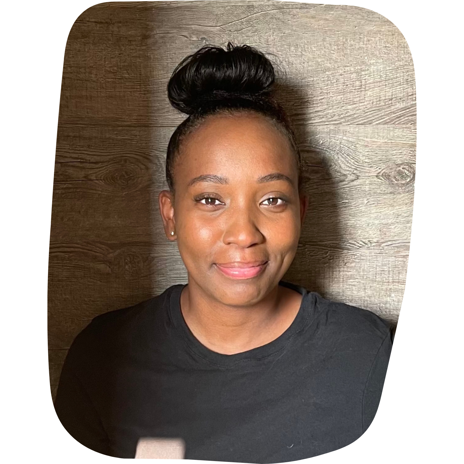 A young woman with a top bun hairstyle, wearing a black shirt, smiling, with a wooden background.