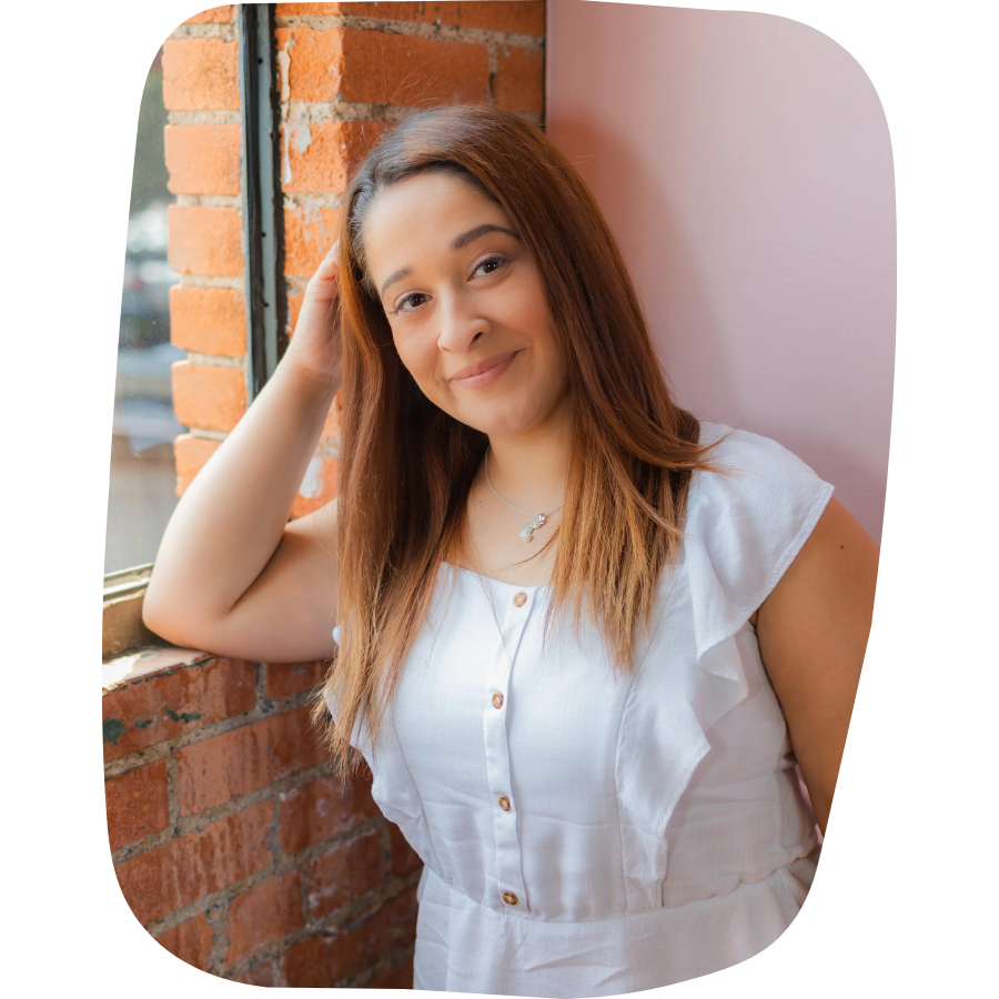 A young woman with long brown hair, wearing a white sleeveless top, standing near a brick wall and window, smiling and looking at the camera.