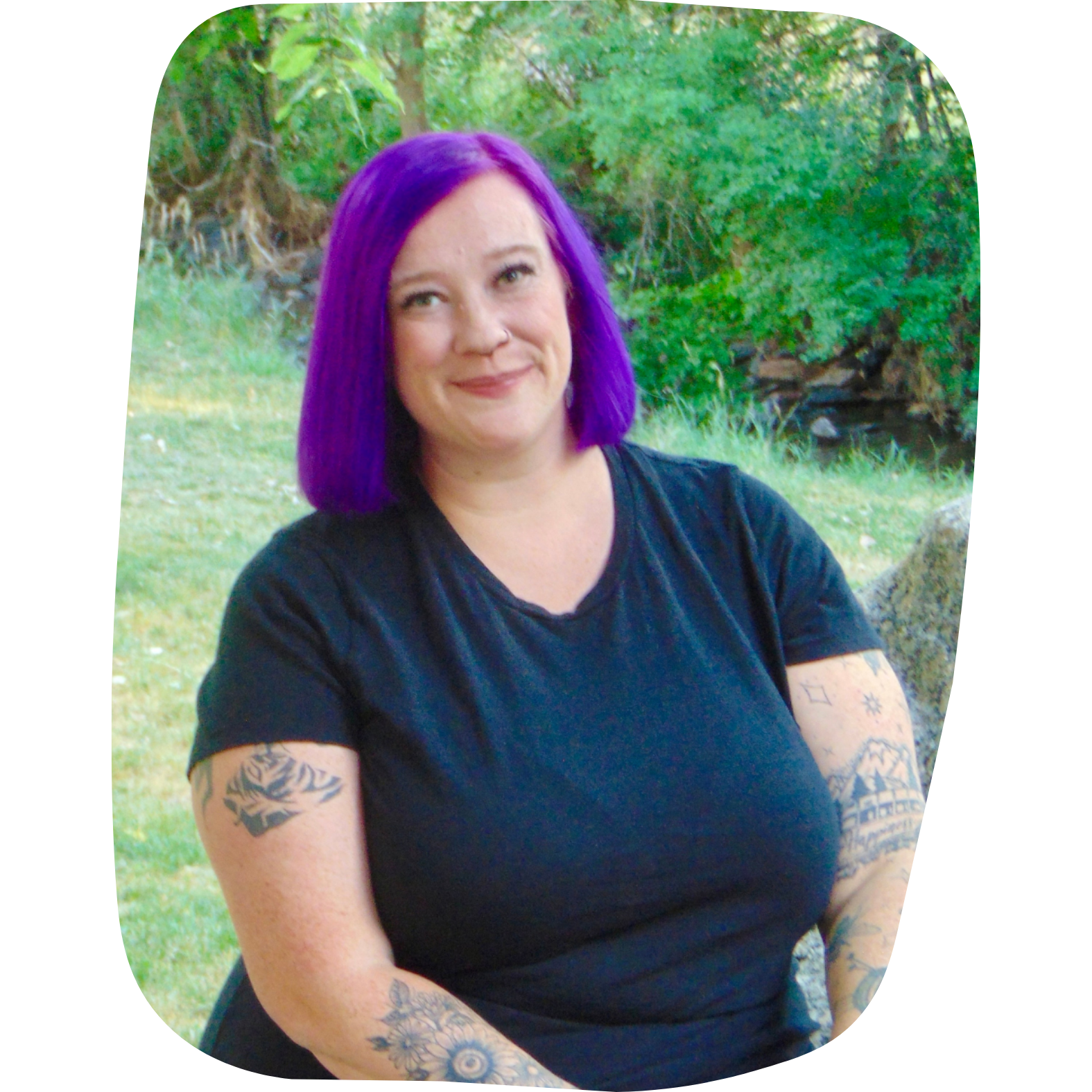 A woman with purple hair wearing a black t-shirt, sitting outdoors near grass and trees, with tattoos on her arms.