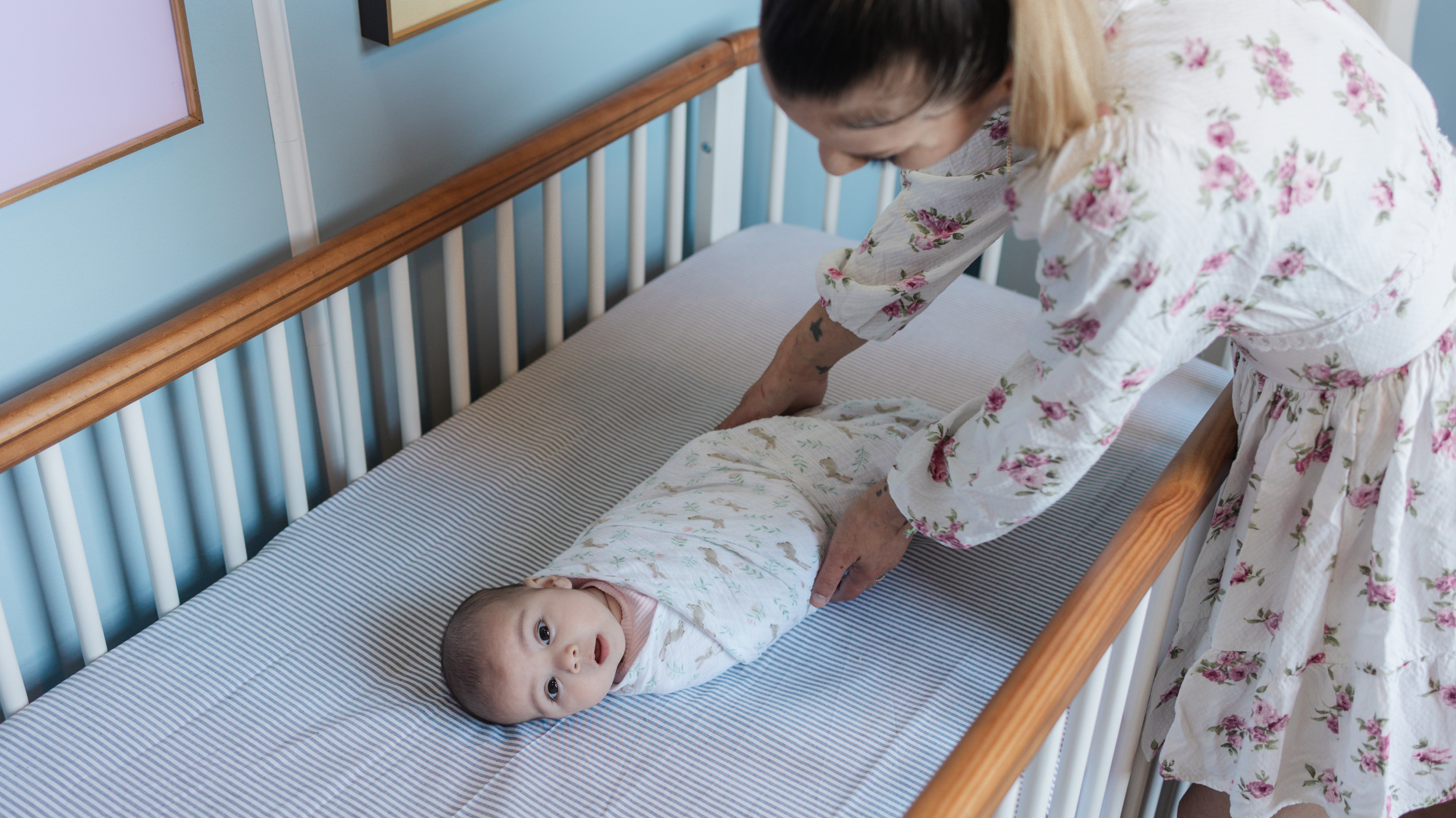 A woman in a floral nightgown adjusting a swaddled baby on a crib mattress.