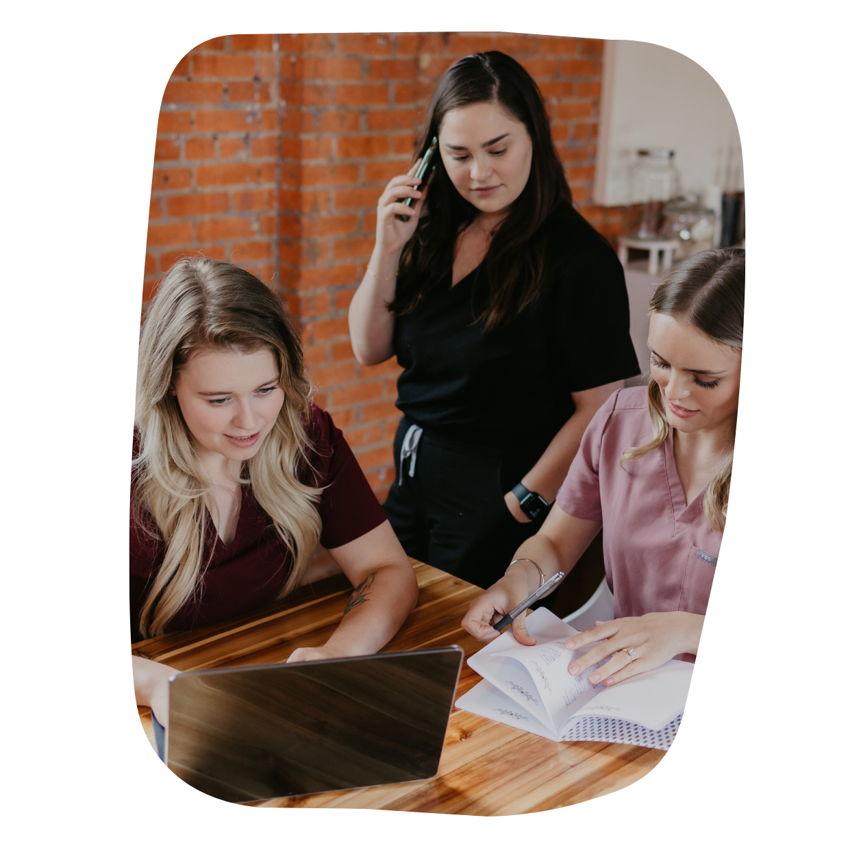 Three women working together around a wooden table with a laptop, notebook, and pen in a room with a brick wall in the background.