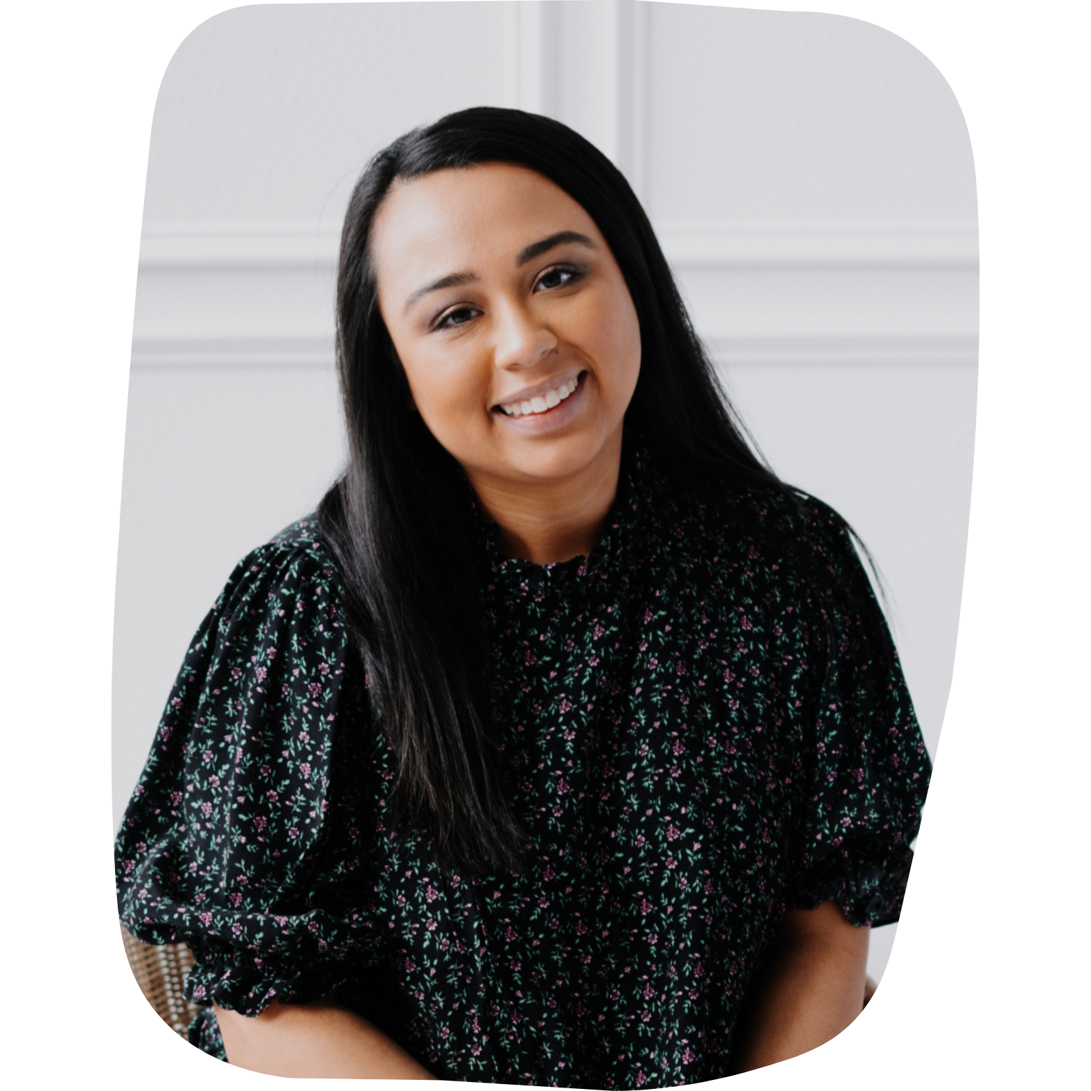 Portrait of a young woman with long black hair, smiling, wearing a black blouse with small colorful floral patterns, sitting indoors with a white wall in the background.