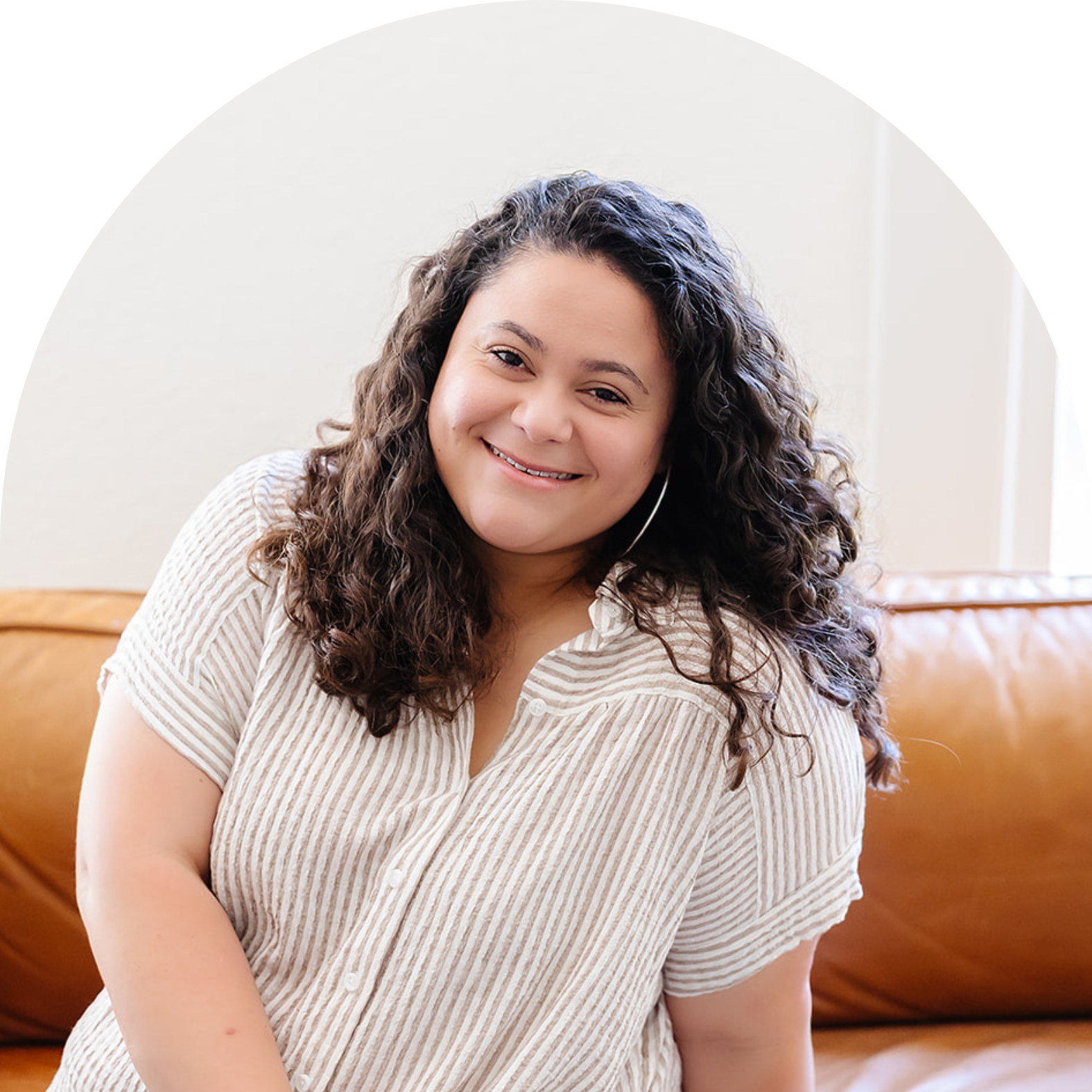 A woman with curly dark hair smiling while sitting on a brown leather couch in a bright room.