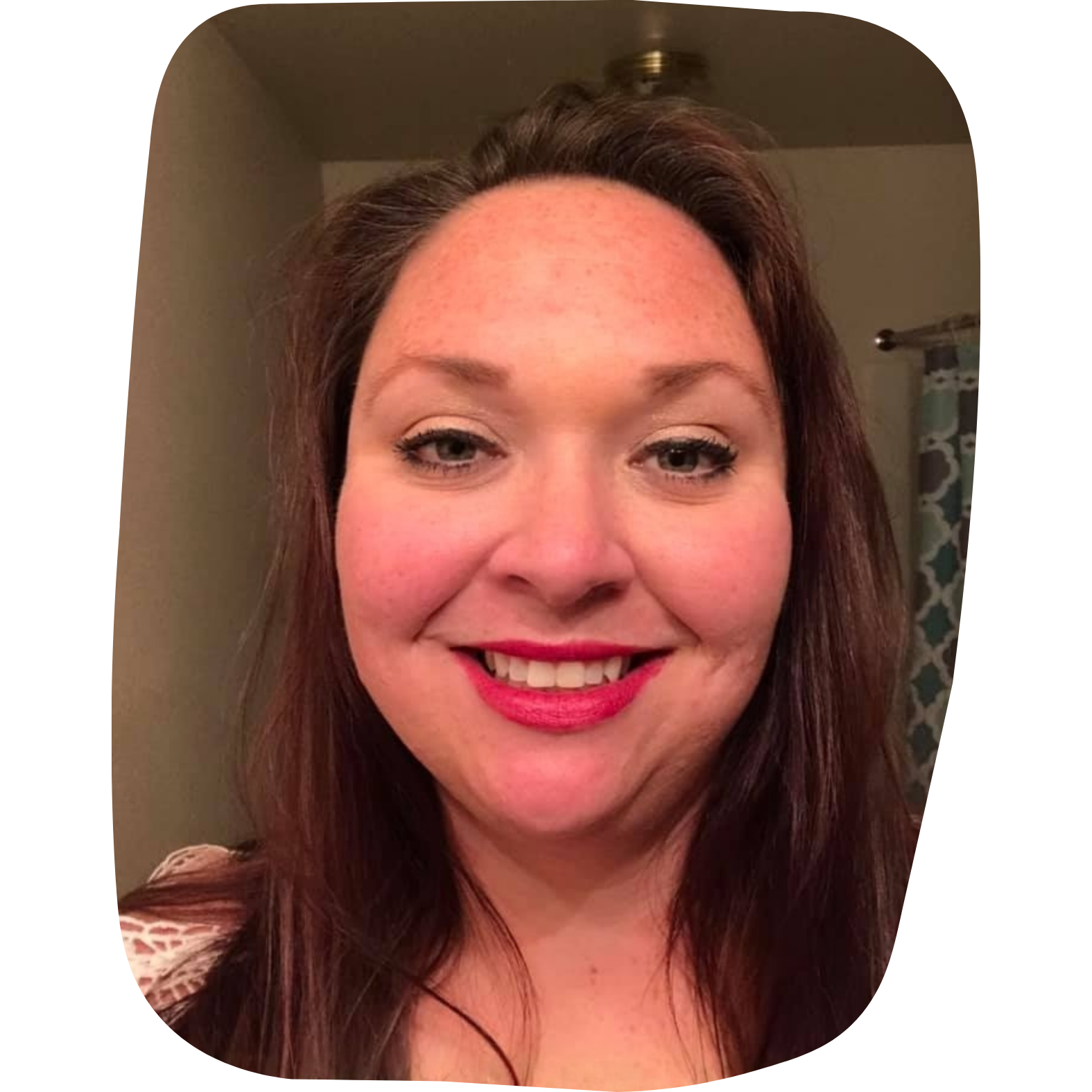 A woman with shoulder-length brown hair smiling, wearing makeup with lipstick, earrings, and a patterned top, in a room with a beige wall and a curtain in the background.