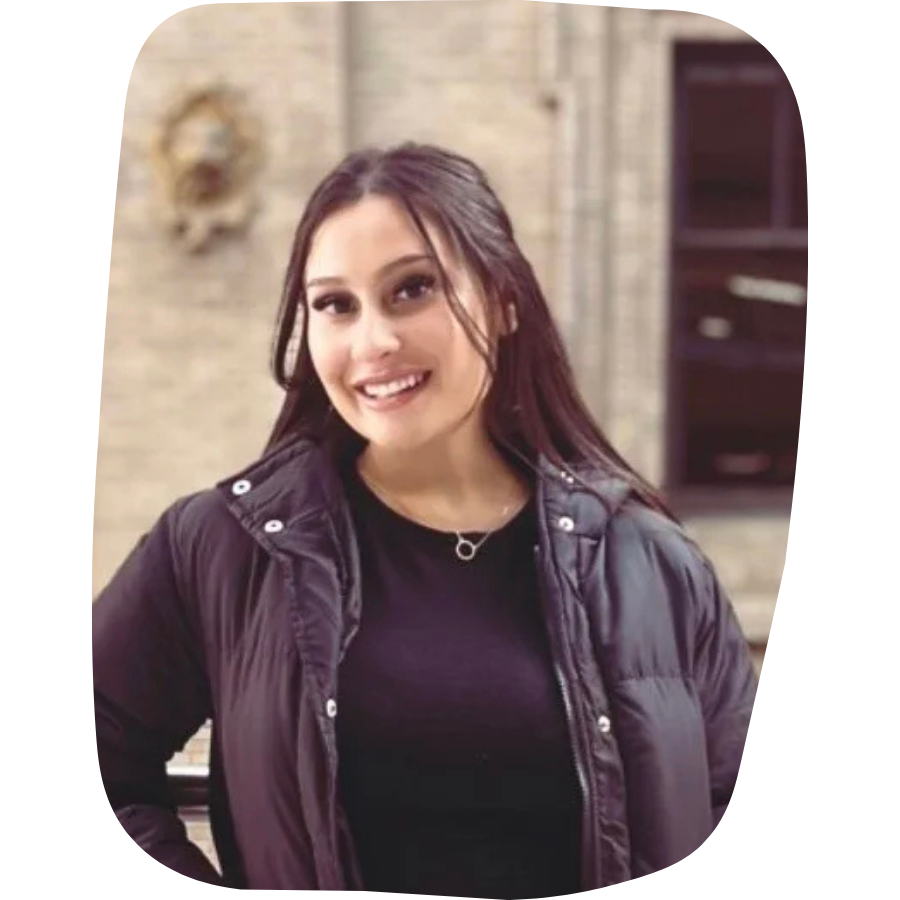 A young woman with dark brown hair, smiling, wearing a black shirt and puffy black jacket, standing indoors in front of a brick wall and a dark bookshelf.