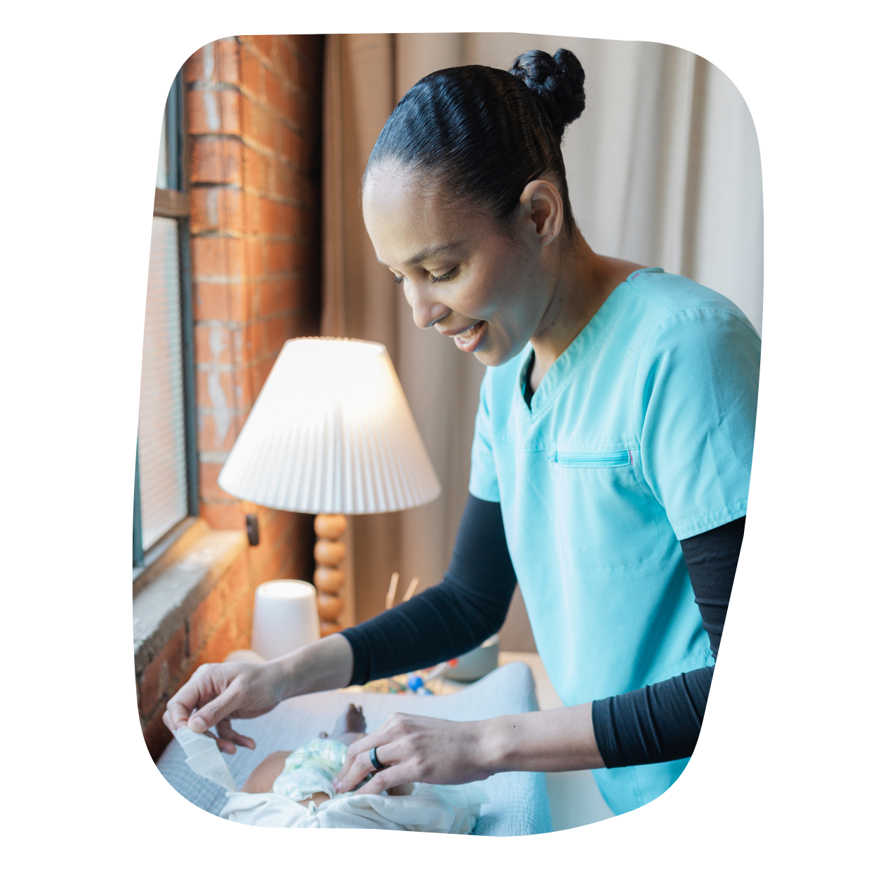 A female healthcare worker in a light blue scrub top caring for a newborn baby in a cozy room with exposed brick wall and a window. She is smiling and gently attending to the baby, who is lying on a changing table.
