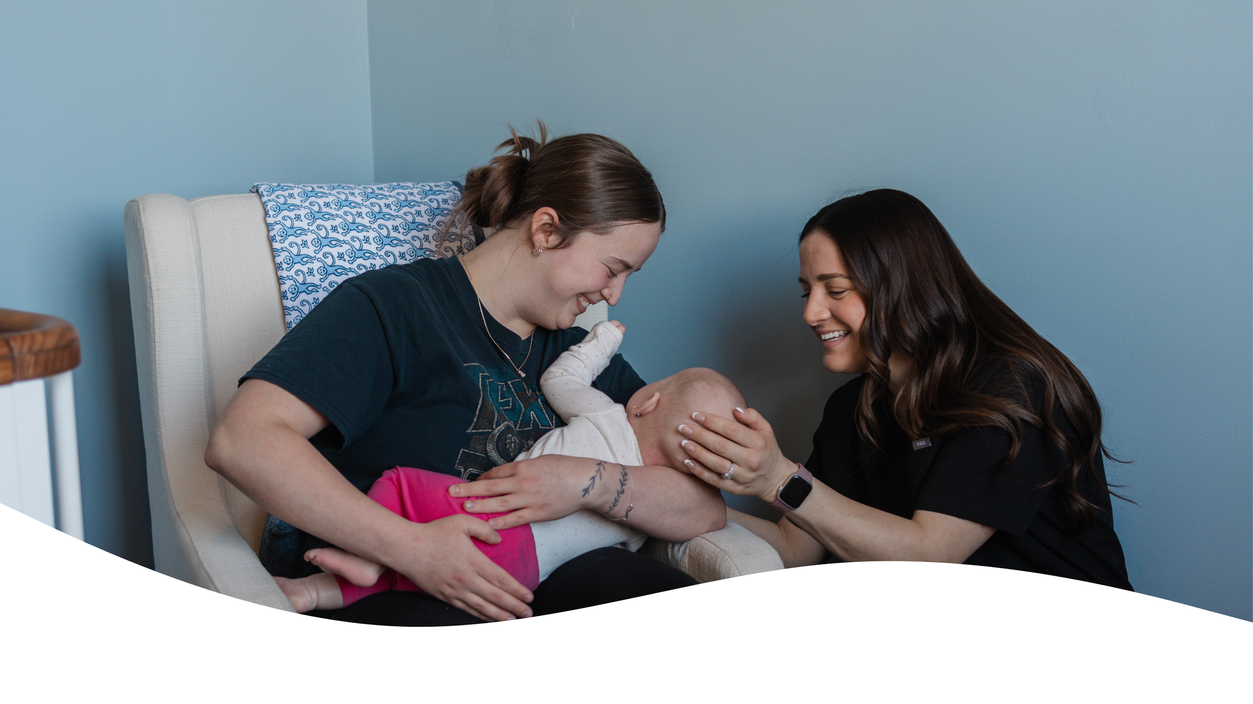 A woman in a black shirt holds a baby's hand while another woman in a dark t-shirt with a Star Wars logo smiles and looks at the baby, sitting in a chair against a light blue wall.