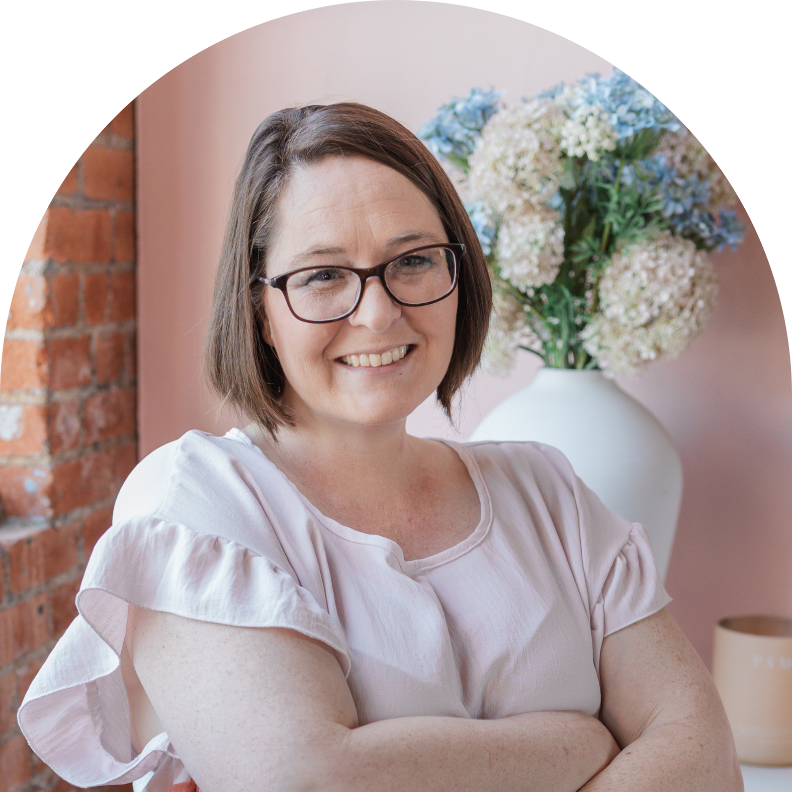 A woman with glasses and short brown hair smiling, in a room with a brick wall and large bouquet of flowers in a white vase in the background.