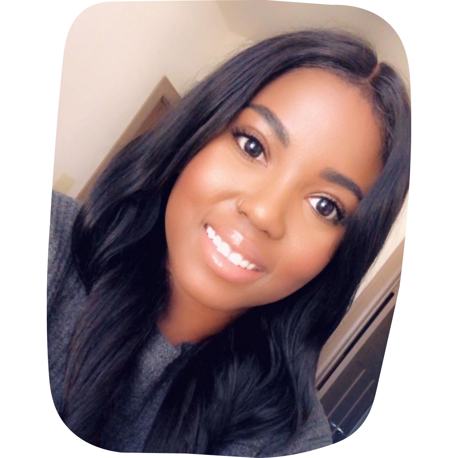Close-up of a smiling woman with long black hair, wearing a gray top and a nose ring, indoors.