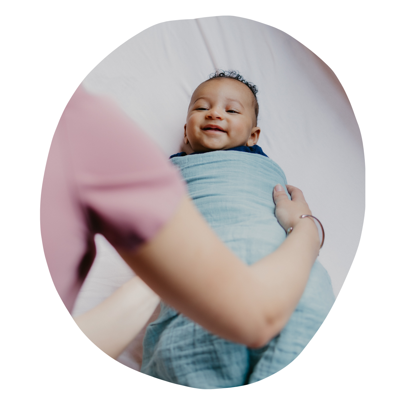 A joyful baby lying on a white surface, being embraced by an adult, with only the adult's arm and part of their pink shirt visible.