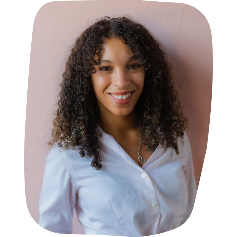 Portrait of a young woman with curly hair, smiling, wearing a white blouse and a silver necklace with a heart-shaped pendant, standing against a pink wall.